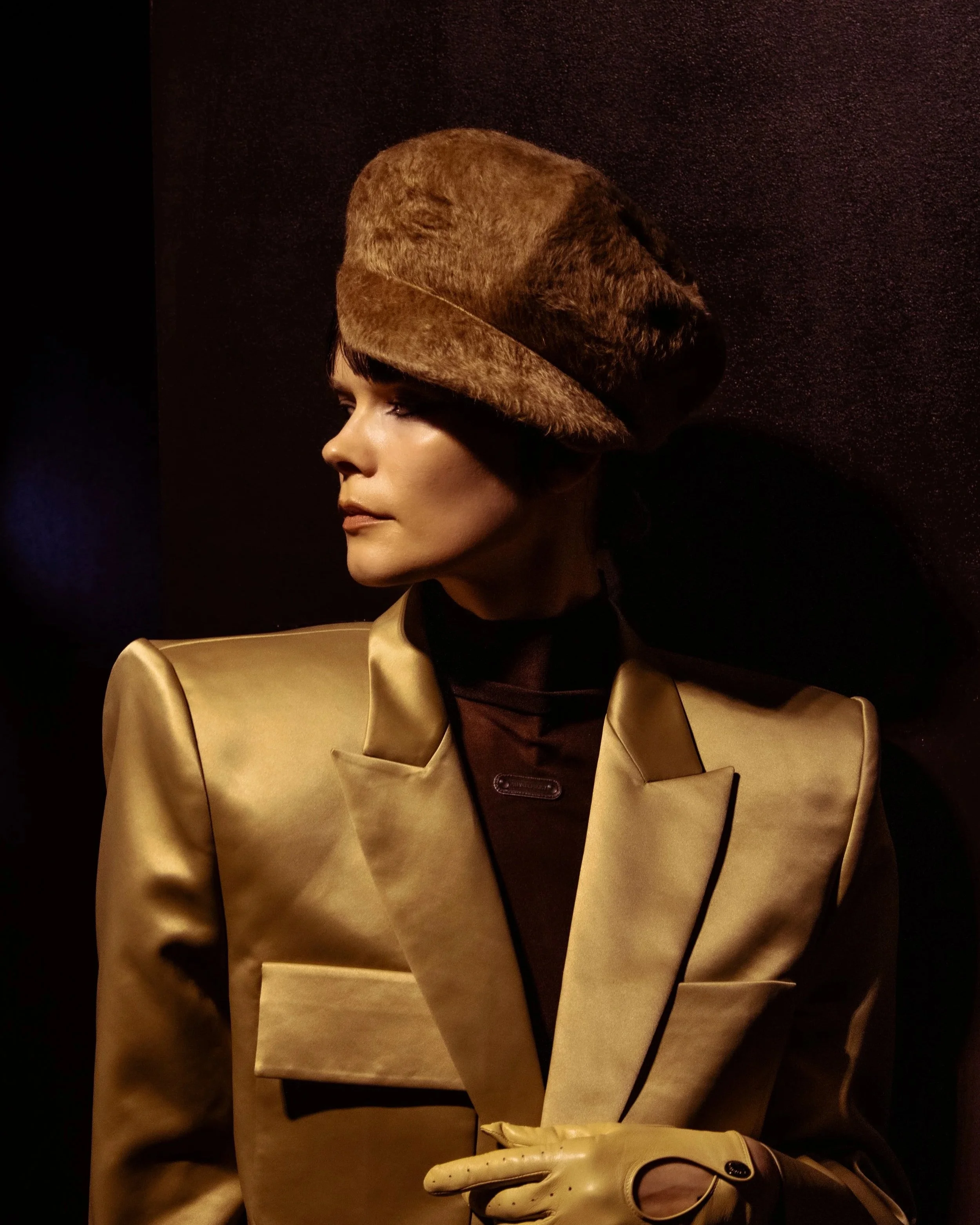 A woman dressed in a gold blazer and beige gloves, wearing a brown fur hat, looking to the side against a dark background.