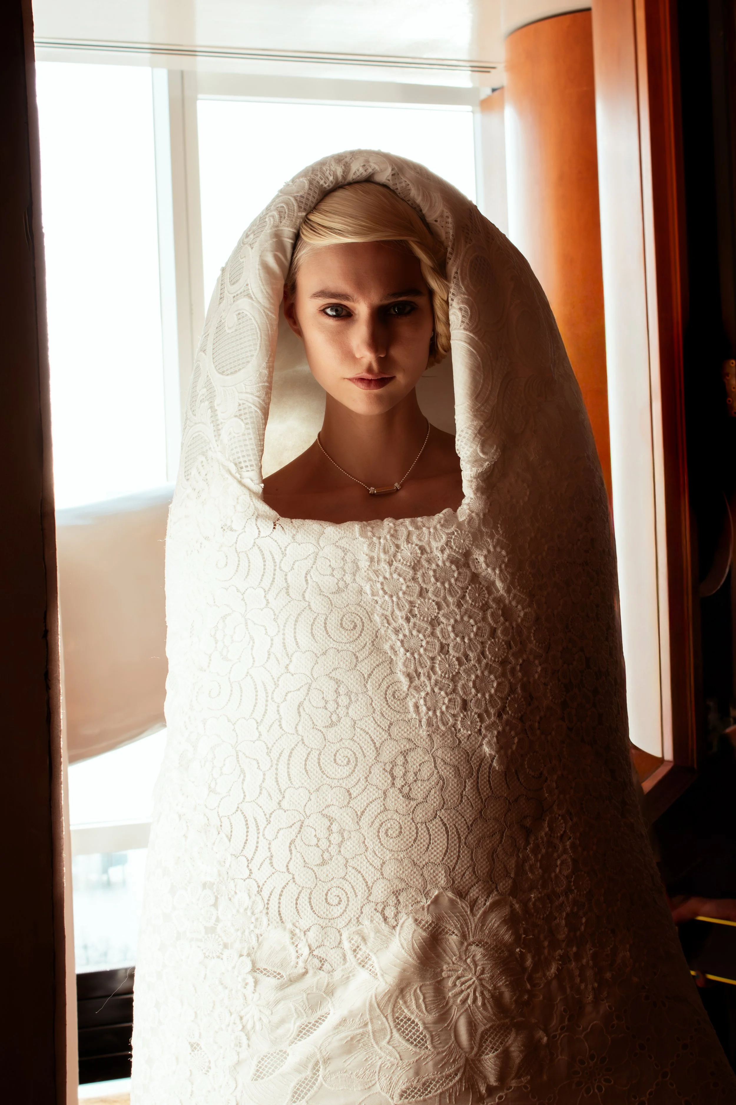 A young woman with short blonde hair looking directly at the camera, standing in front of a window, wrapped in a white lace fabric that drapes over her head and body.