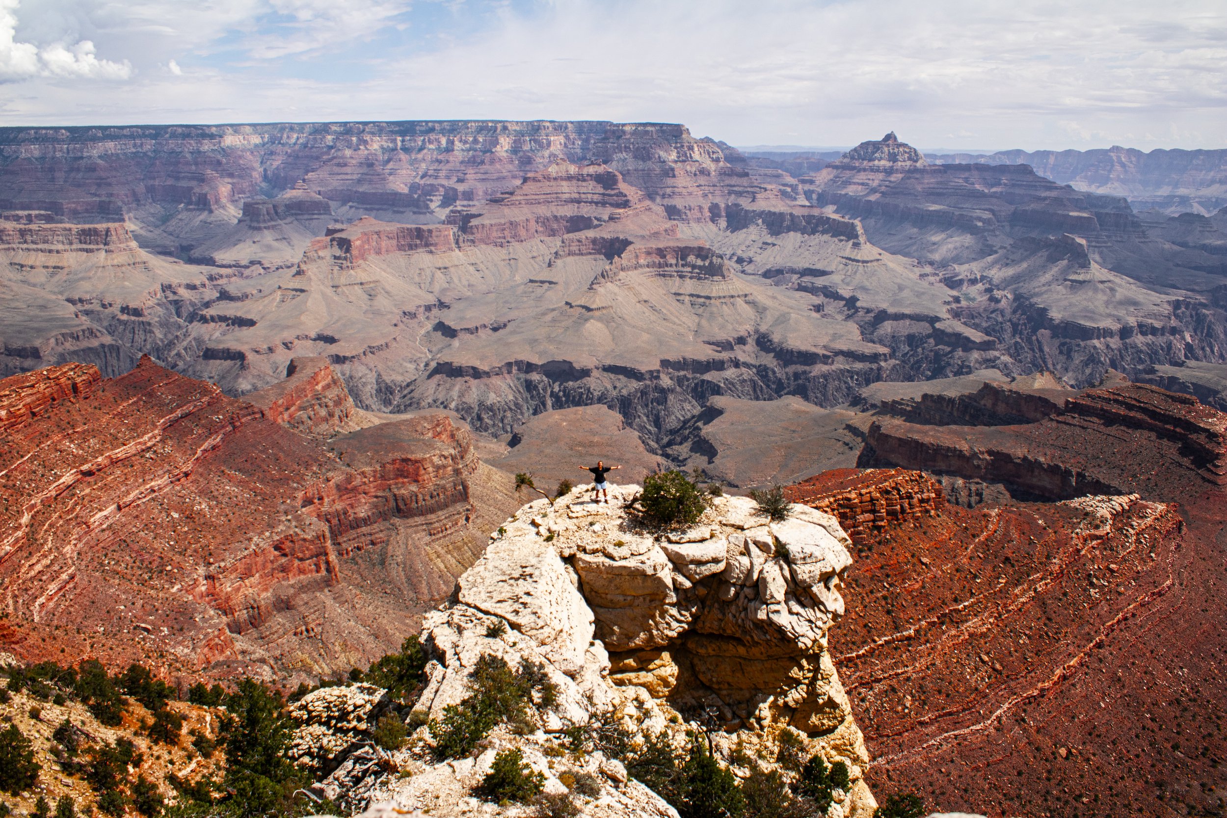 Person standing with arms outstretched on a rocky ledge overlooking the Grand Canyon with layered red, orange, and brown cliffs and a cloudy sky.