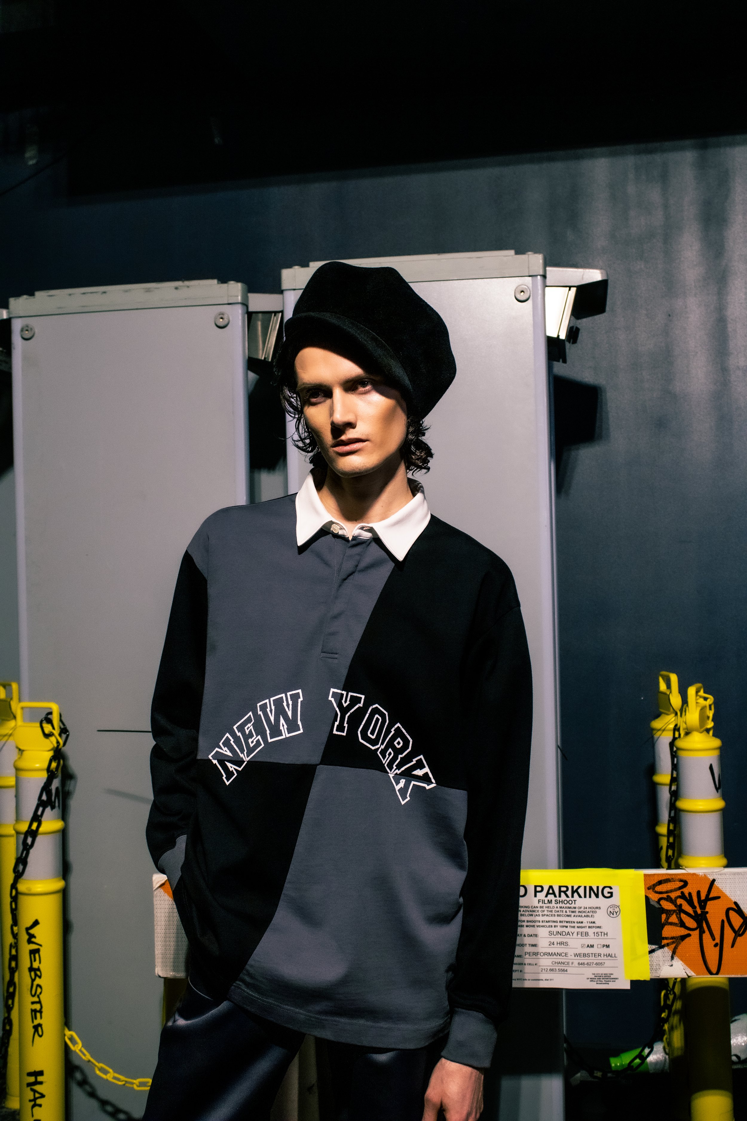 Young man with dark, wavy hair wearing a black beret and a bomber jacket with 'New York' text, standing in front of yellow barricades.