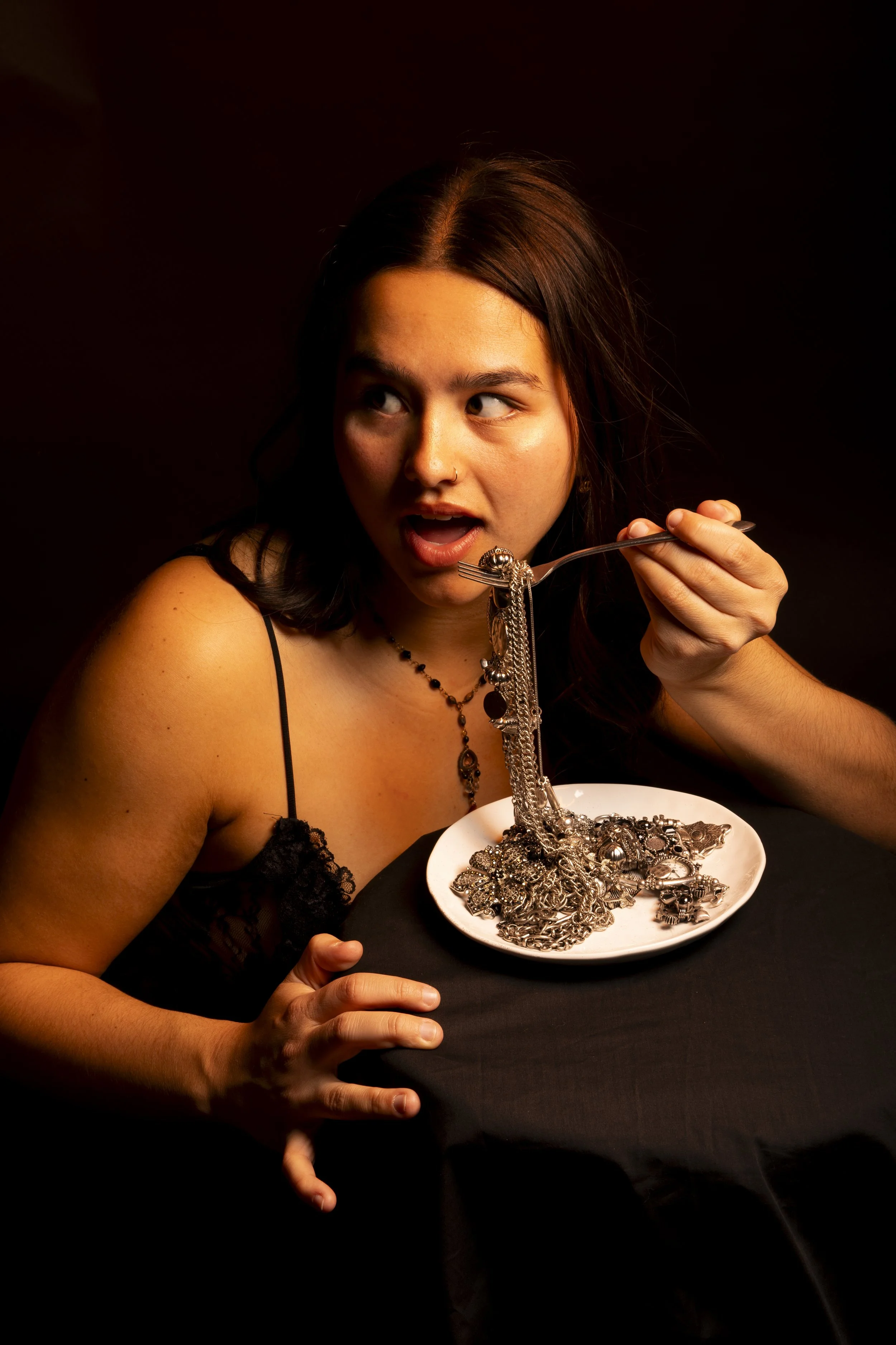 A woman in a black dress with spaghetti straps and necklace is holding a fork with tangled jewelry over a white plate filled with jewelry, on a black background.