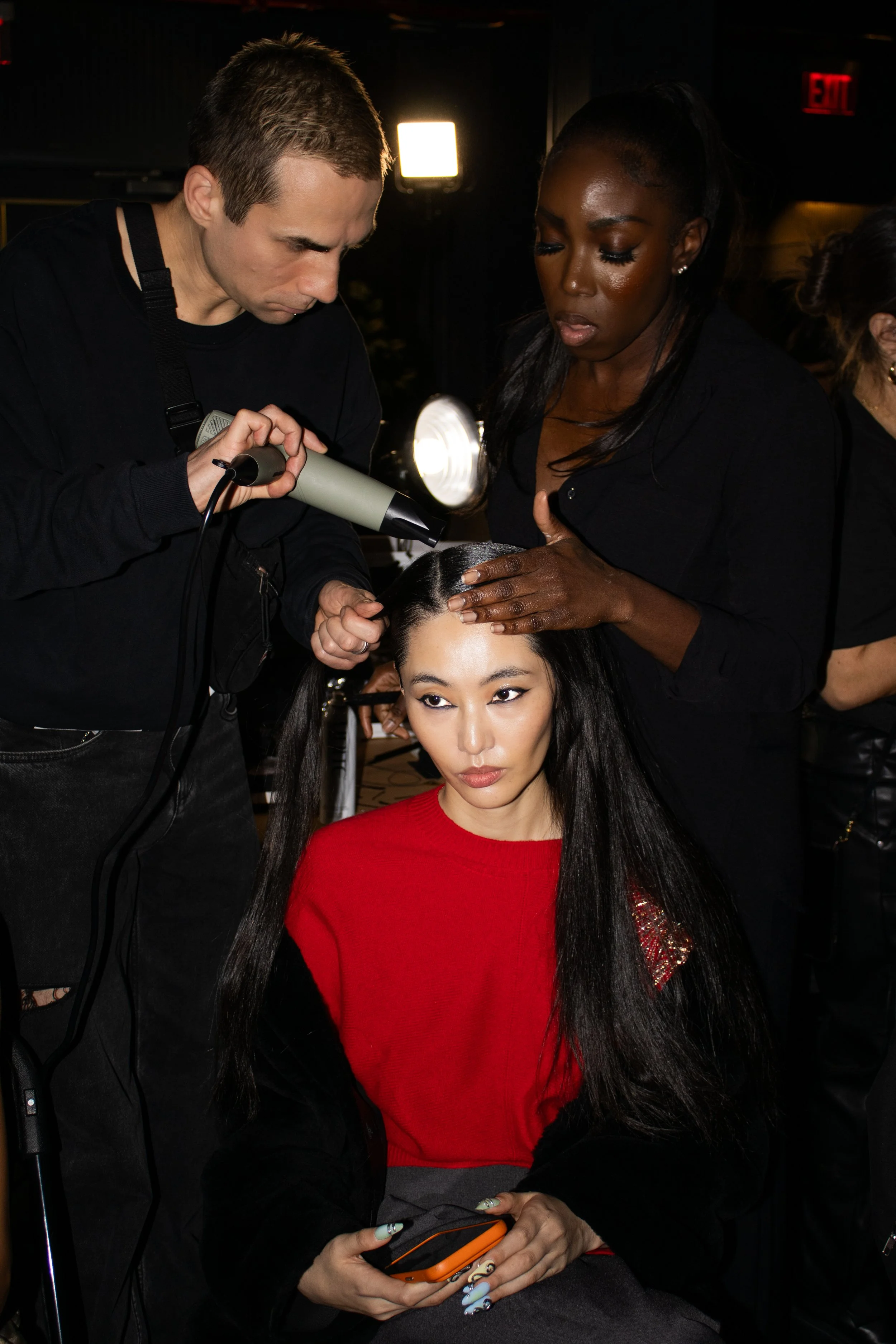 A woman getting her hair styled with a hairdryer while holding her phone, with a stylist drying and preparing her hair in a salon setting.