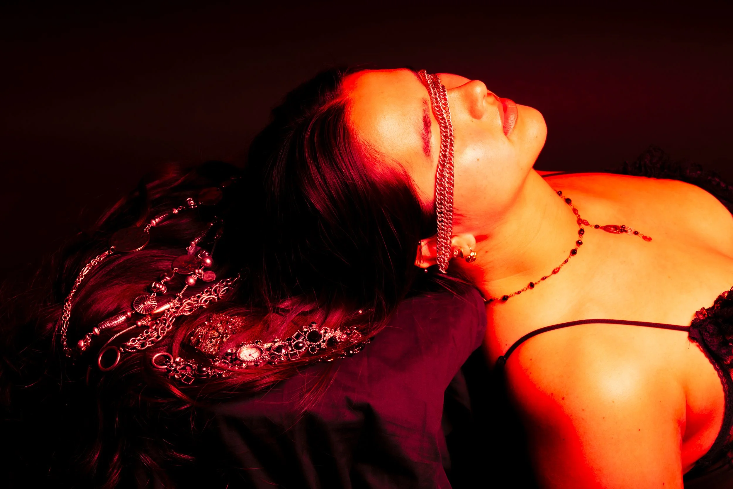 A woman lying down, illuminated by red light, wearing jewelry including chains, earrings, and a choker, dressed in dark clothing with a black lace top.