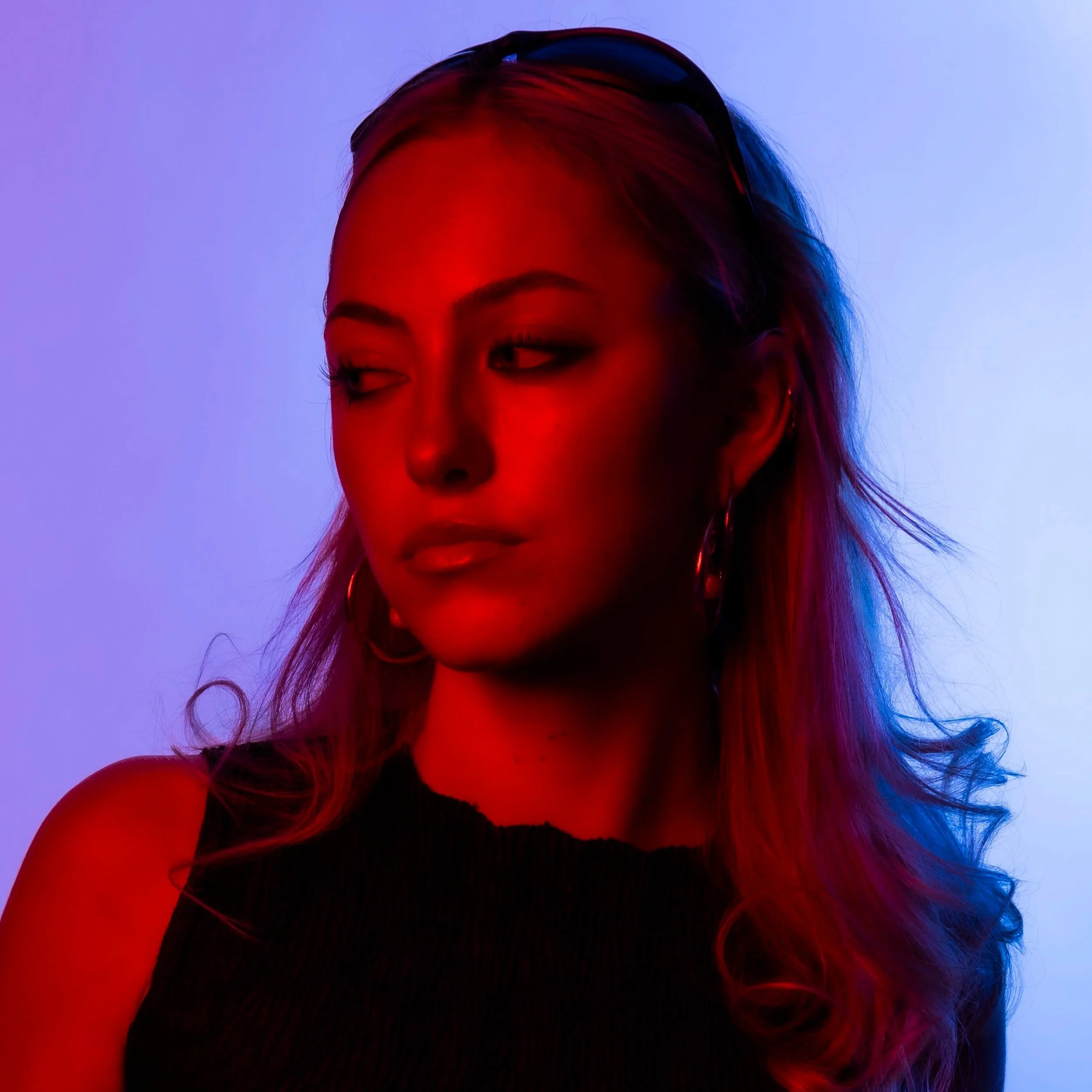 A woman with long hair, hoop earrings, and sunglasses on her head, looking to the side, illuminated with red and blue lighting.