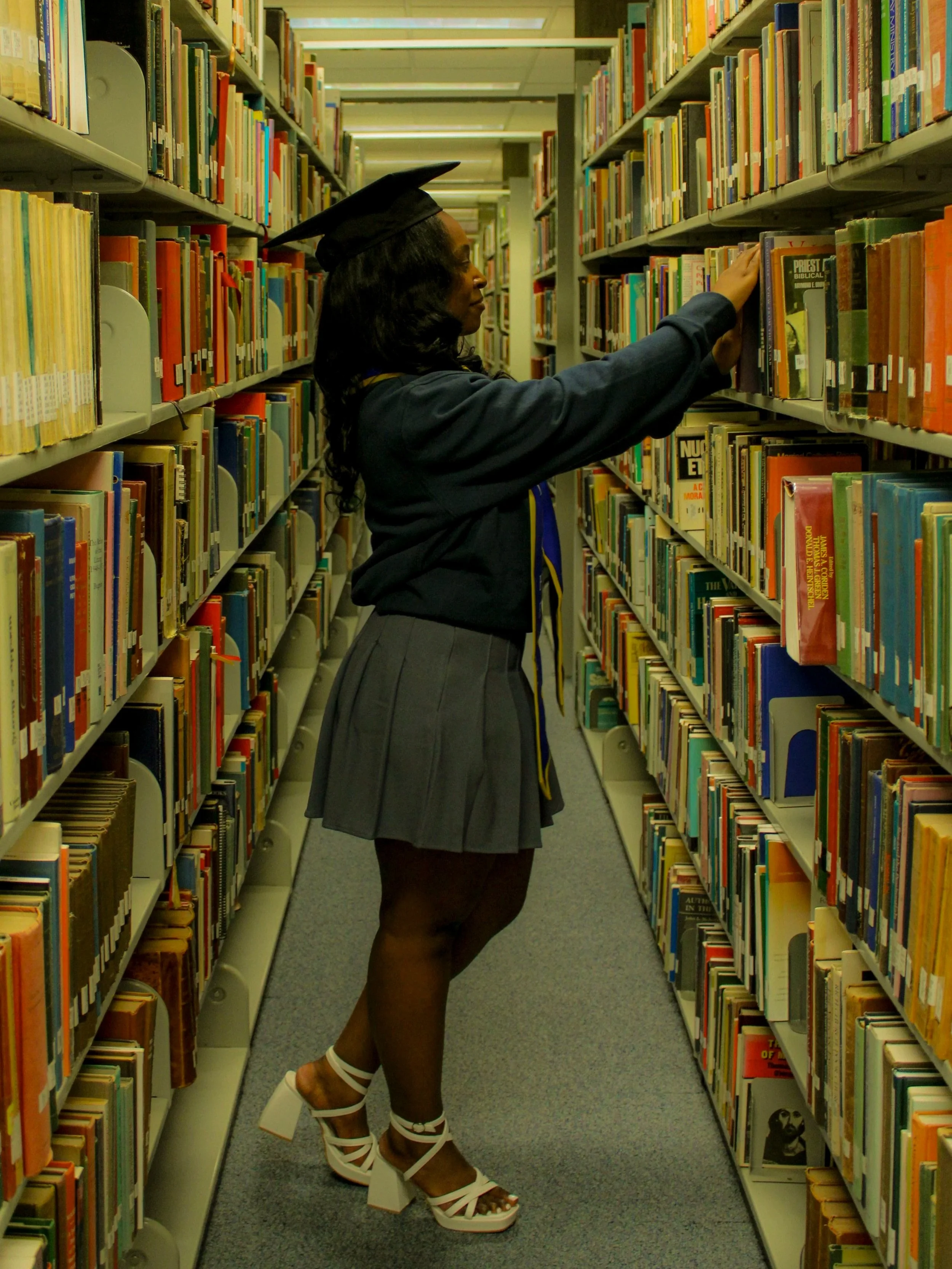 A woman in a graduation cap and gown standing in a library aisle, selecting a book from the shelf.