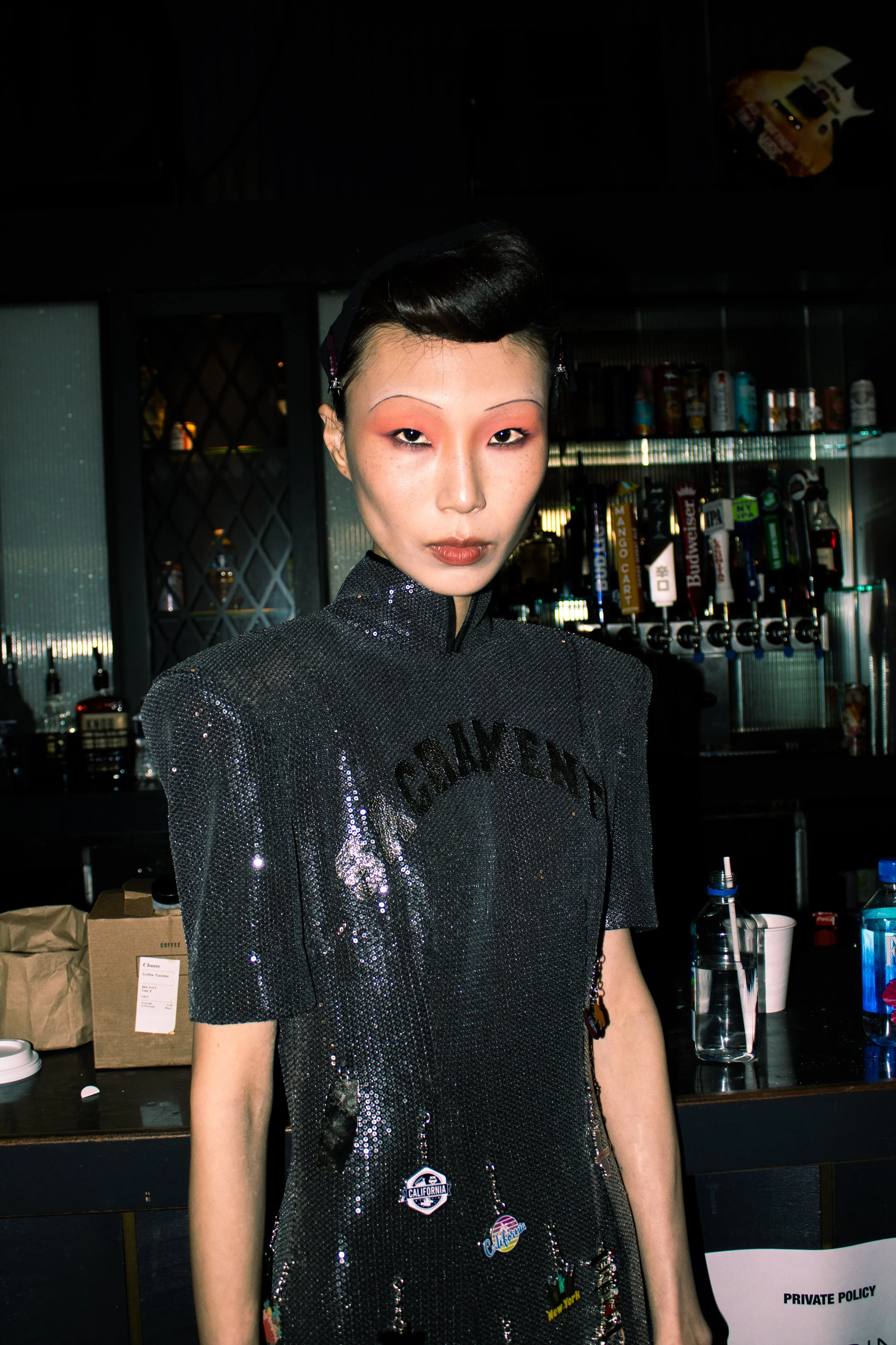A woman with short black hair and makeup standing in a bar area with liquor bottles and a drink dispenser in the background.