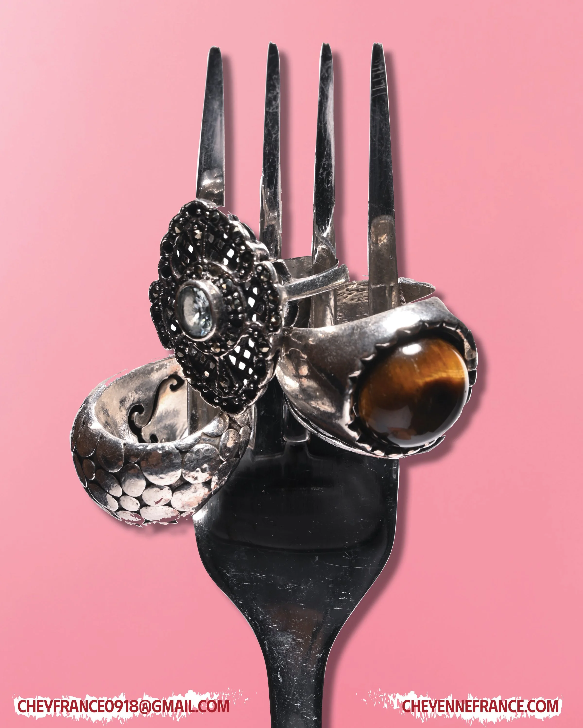 A fork holding three rings with decorative designs and gemstones, set against a pink background.