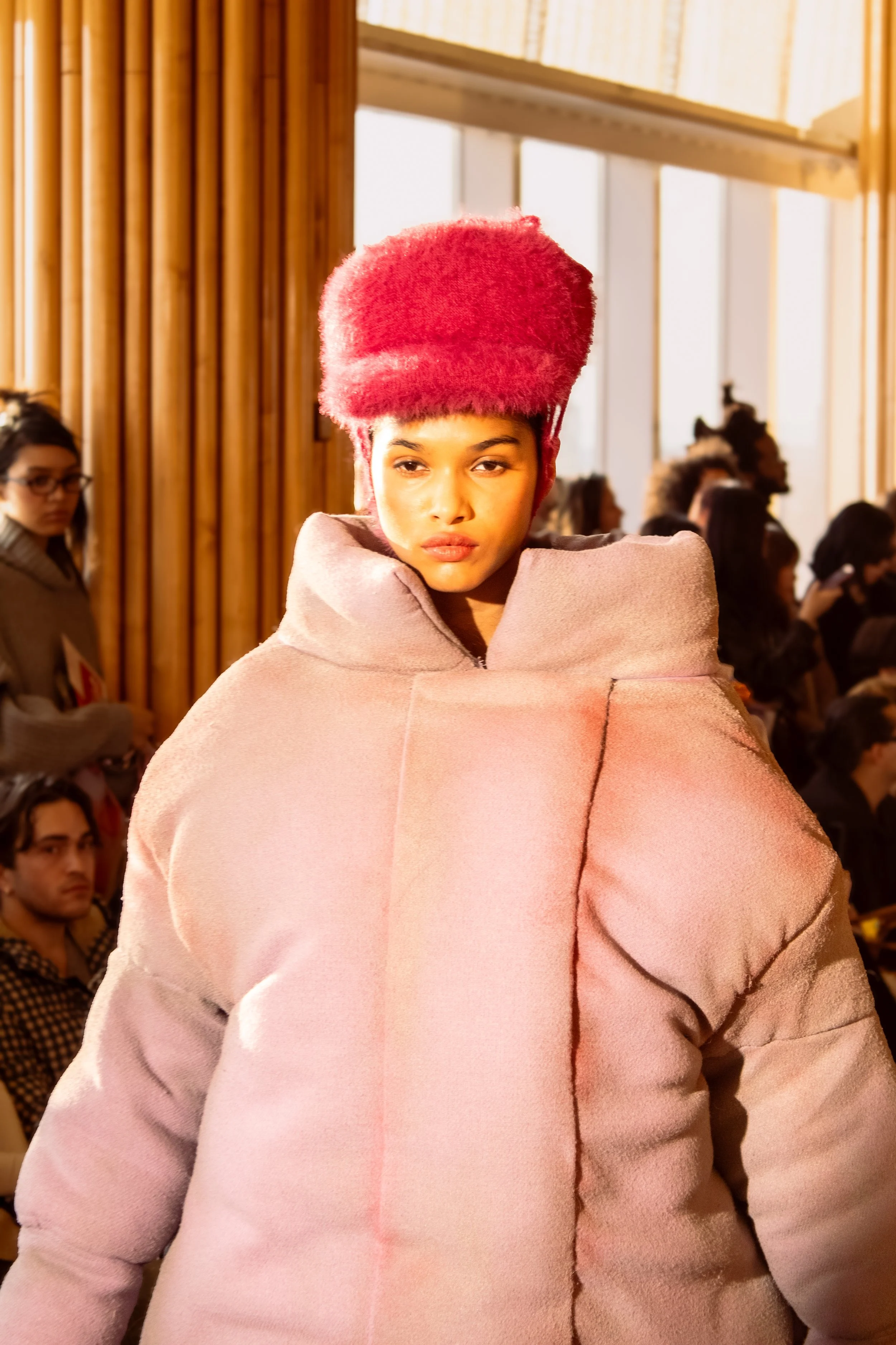 Fashion model on runway wearing a light purple puffer coat and a bright pink furry hat, with an audience in the background.