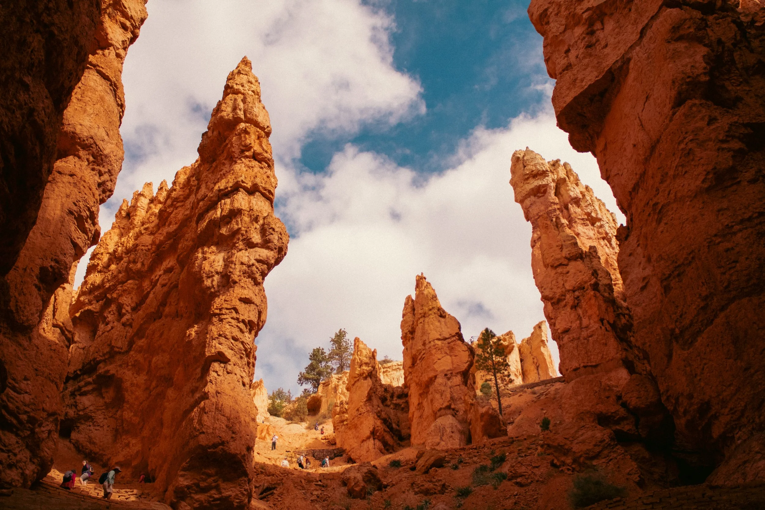 View of tall orange rock formations in a desert landscape with blue sky and clouds, with people hiking at the base.