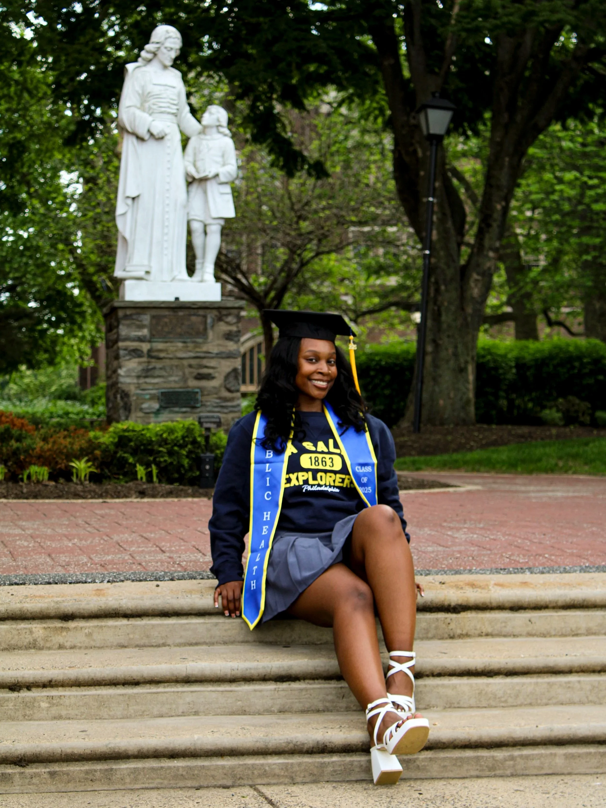 A young woman in graduation attire, including a cap and gown with a sash, sitting on steps with a smile. In the background, there is a statue of a woman and a child, trees, and a lamp post in a park-like setting.