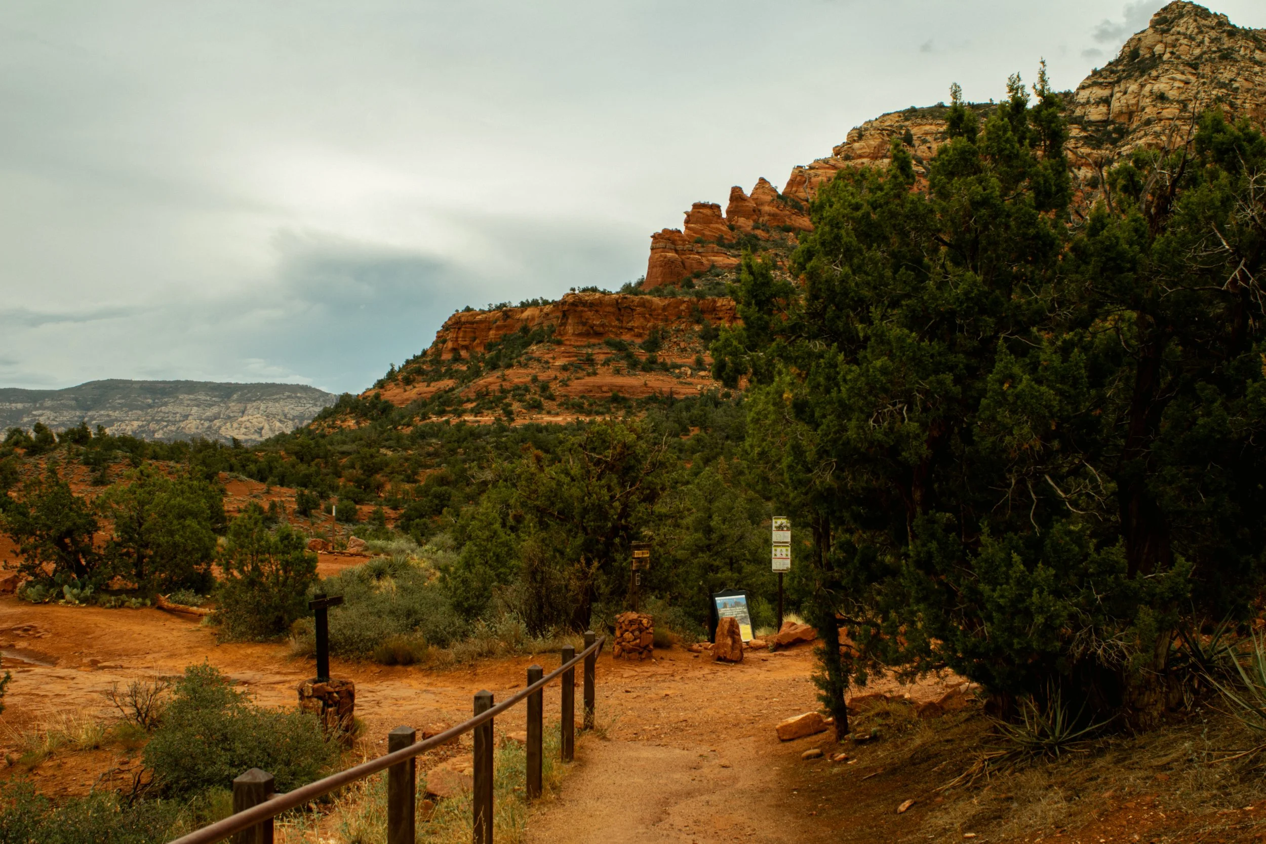 A dirt trail in a desert landscape with green trees, red rocky hills, and mountains in the background.