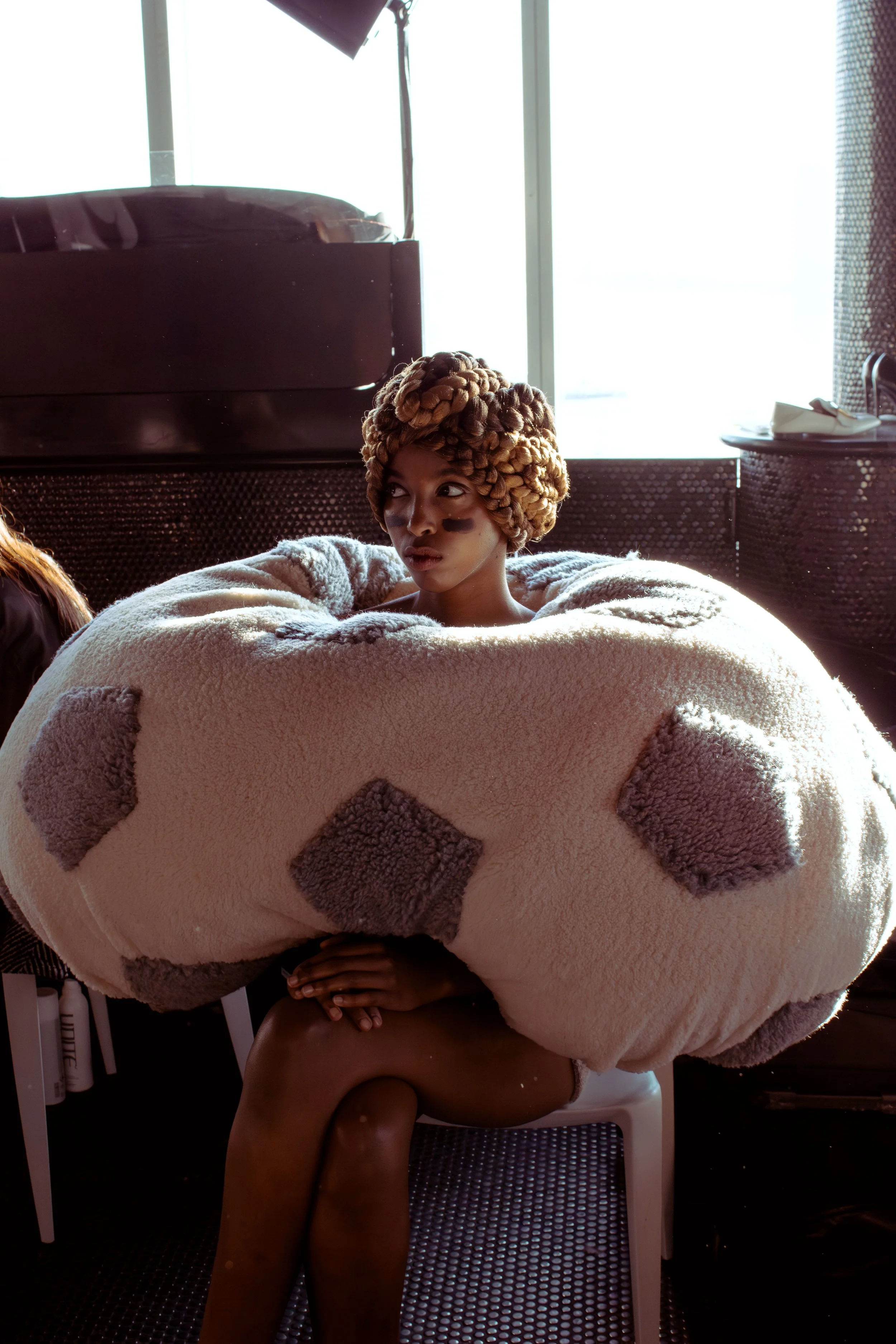 A young girl dressed as a soccer ball, with braided hair, sitting on a white chair in a room with large windows and sunlight.