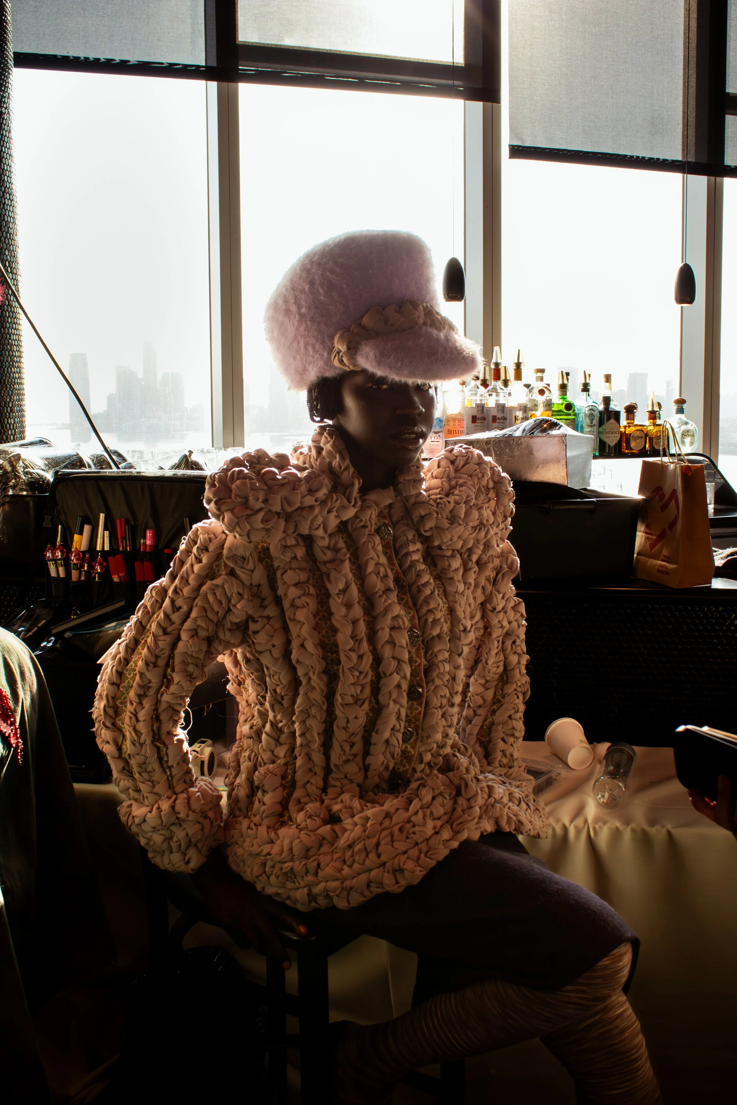 A person sitting in a room with large windows, wearing a fluffy pink hat and a textured purple jacket with exaggerated shoulders, with a city skyline visible in the background.