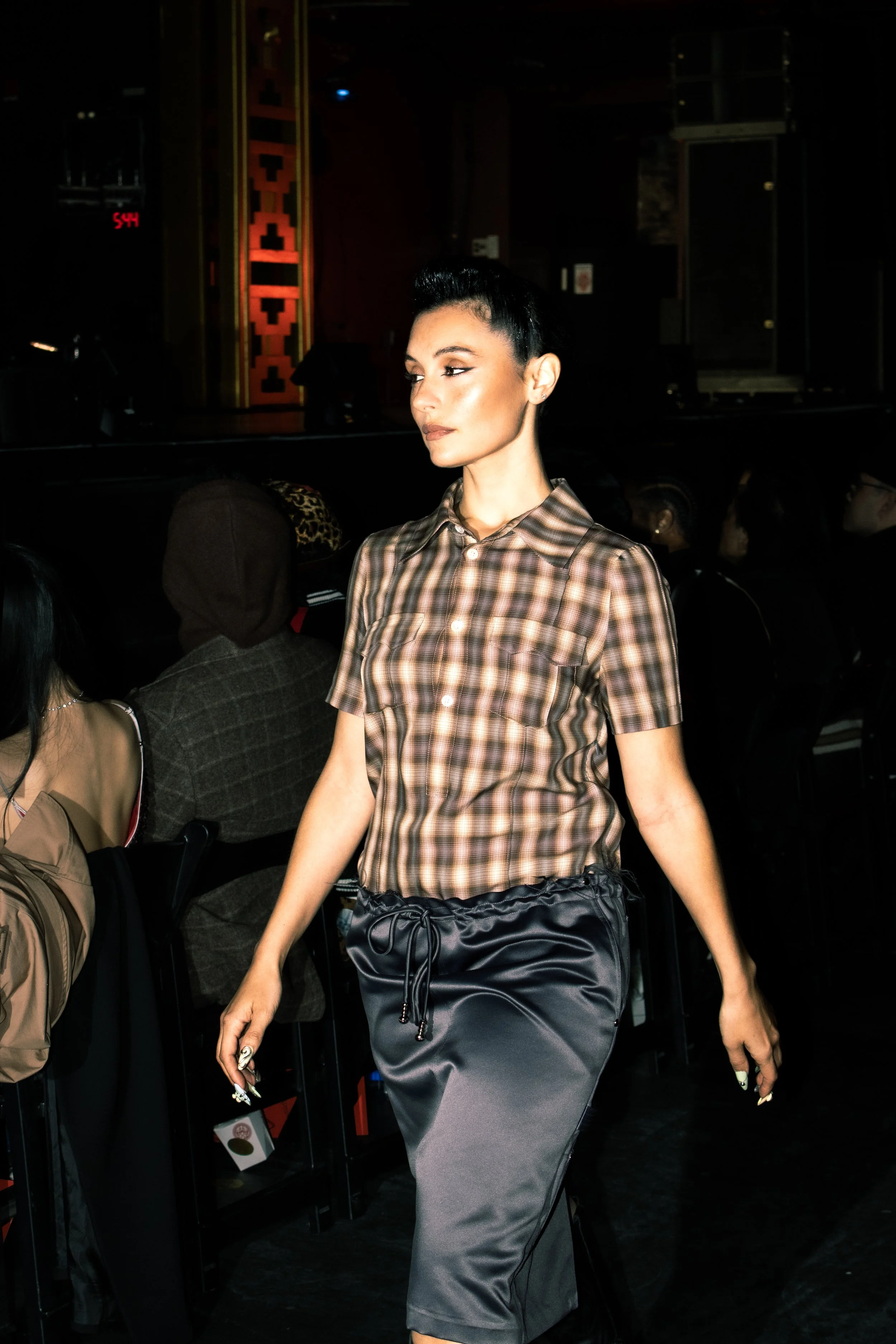 A woman with dark hair styled in a bun, wearing a short-sleeved plaid shirt and what appears to be a satin skirt, walking in an indoor venue with dark background and seated audience.