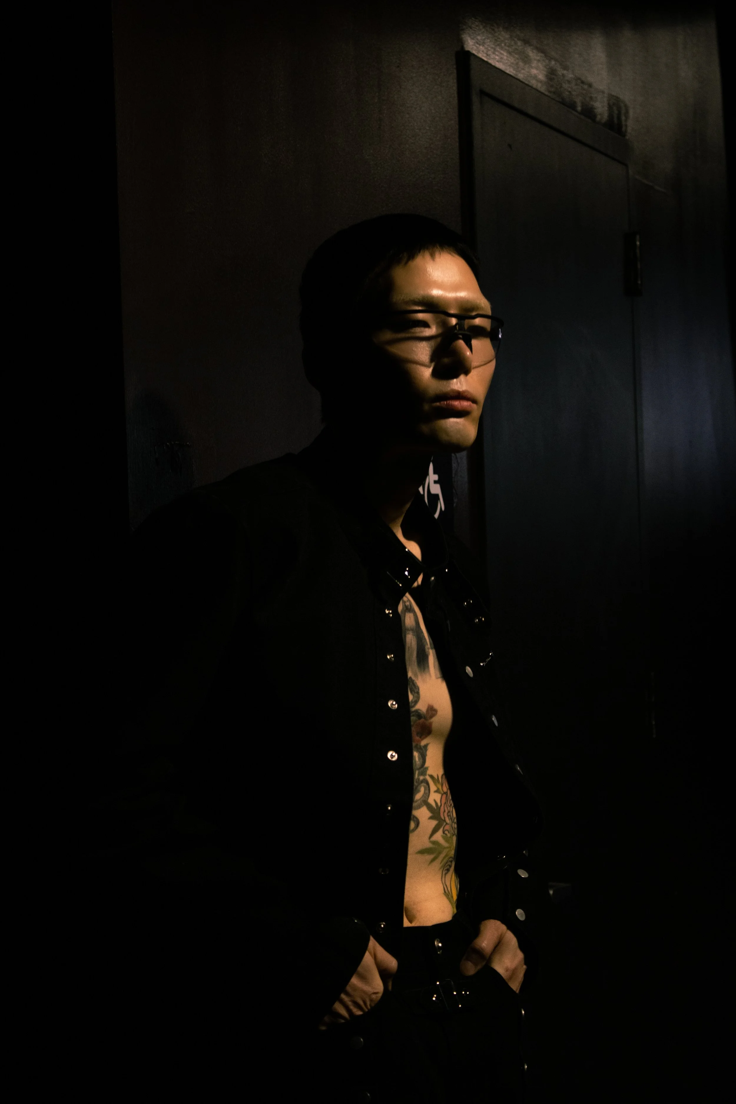 A person with tattoos, glasses, and black clothing standing in a dark room with minimal lighting.