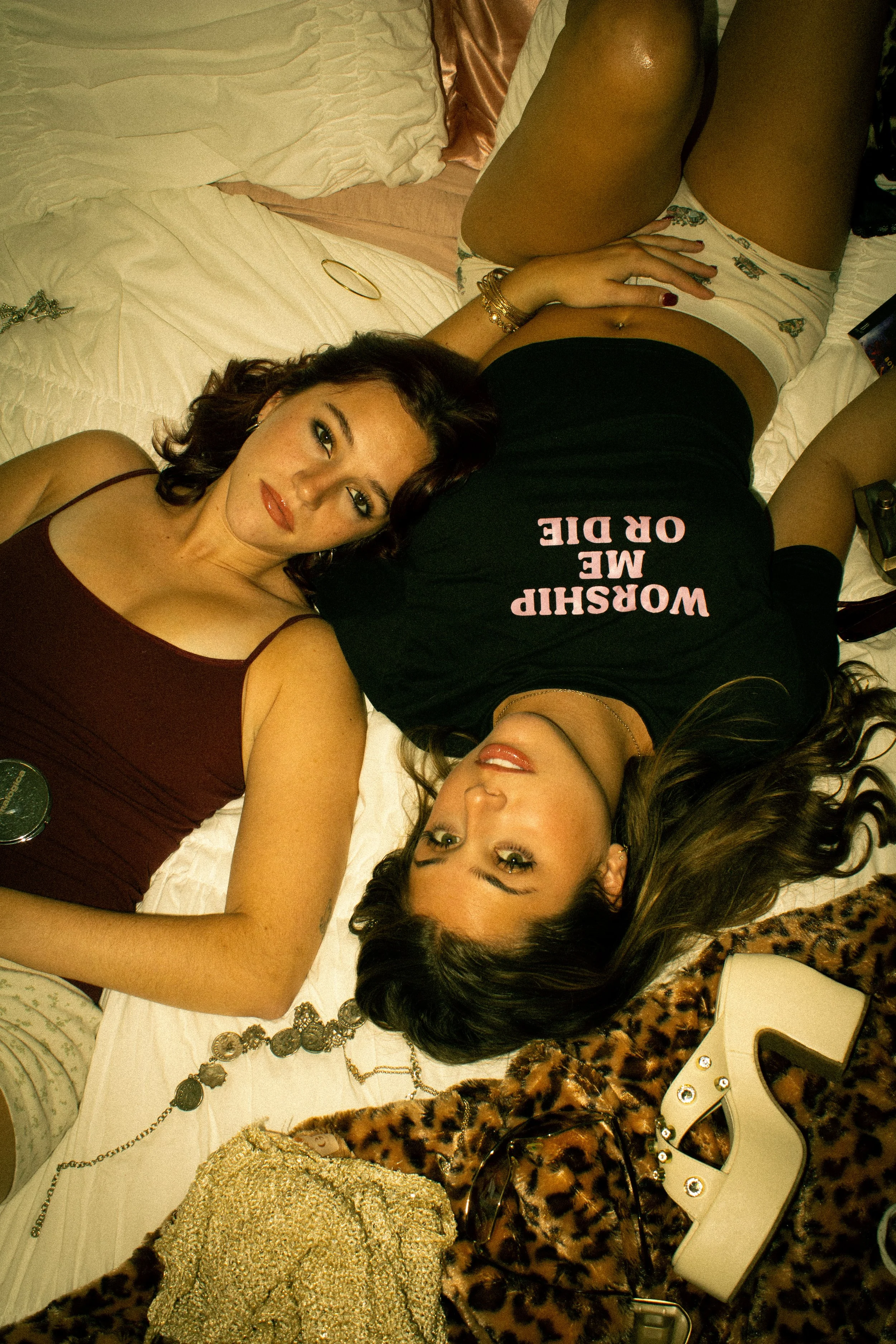 Two women lying on a bed, one facing up and the other facing down. One woman has dark hair and is wearing a maroon top with spaghetti straps, while the other has lighter hair and is wearing a black t-shirt with text that reads 'WORSHIP ME OR DIE.' Th