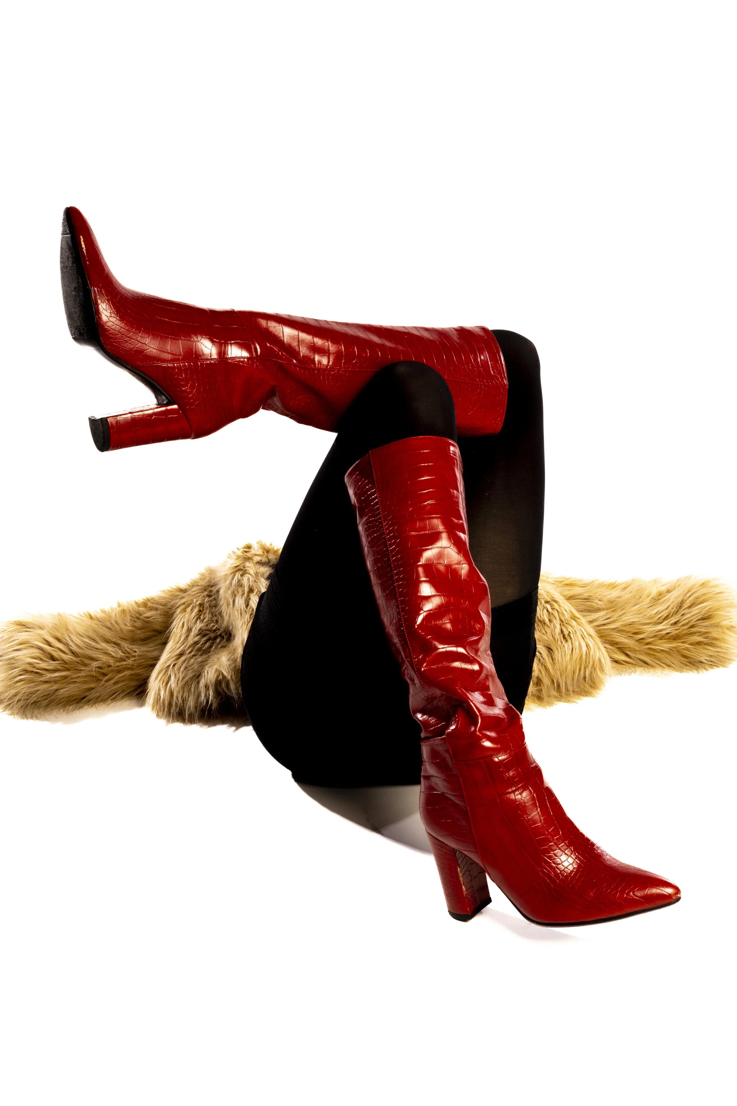 A woman wearing red high-heeled boots with a crocodile pattern, black stockings, and a tan furry coat, against a white background