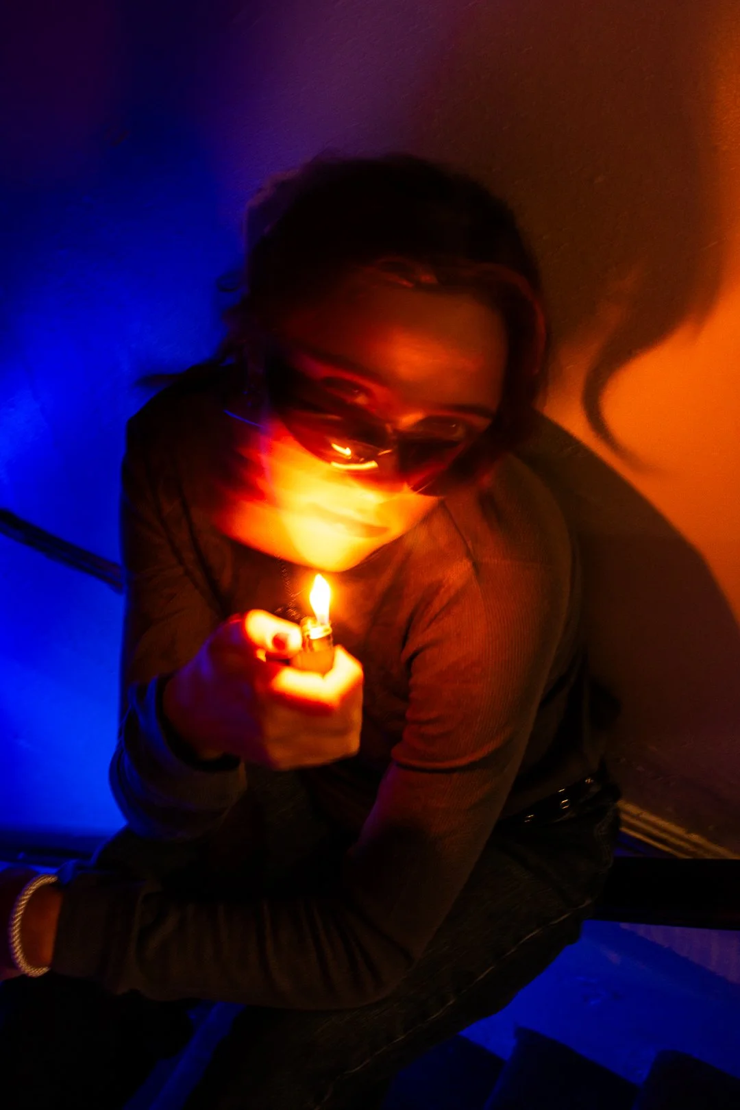 Person with long hair wearing sunglasses, holding a lit lighter in a dark room with blue and orange lighting.
