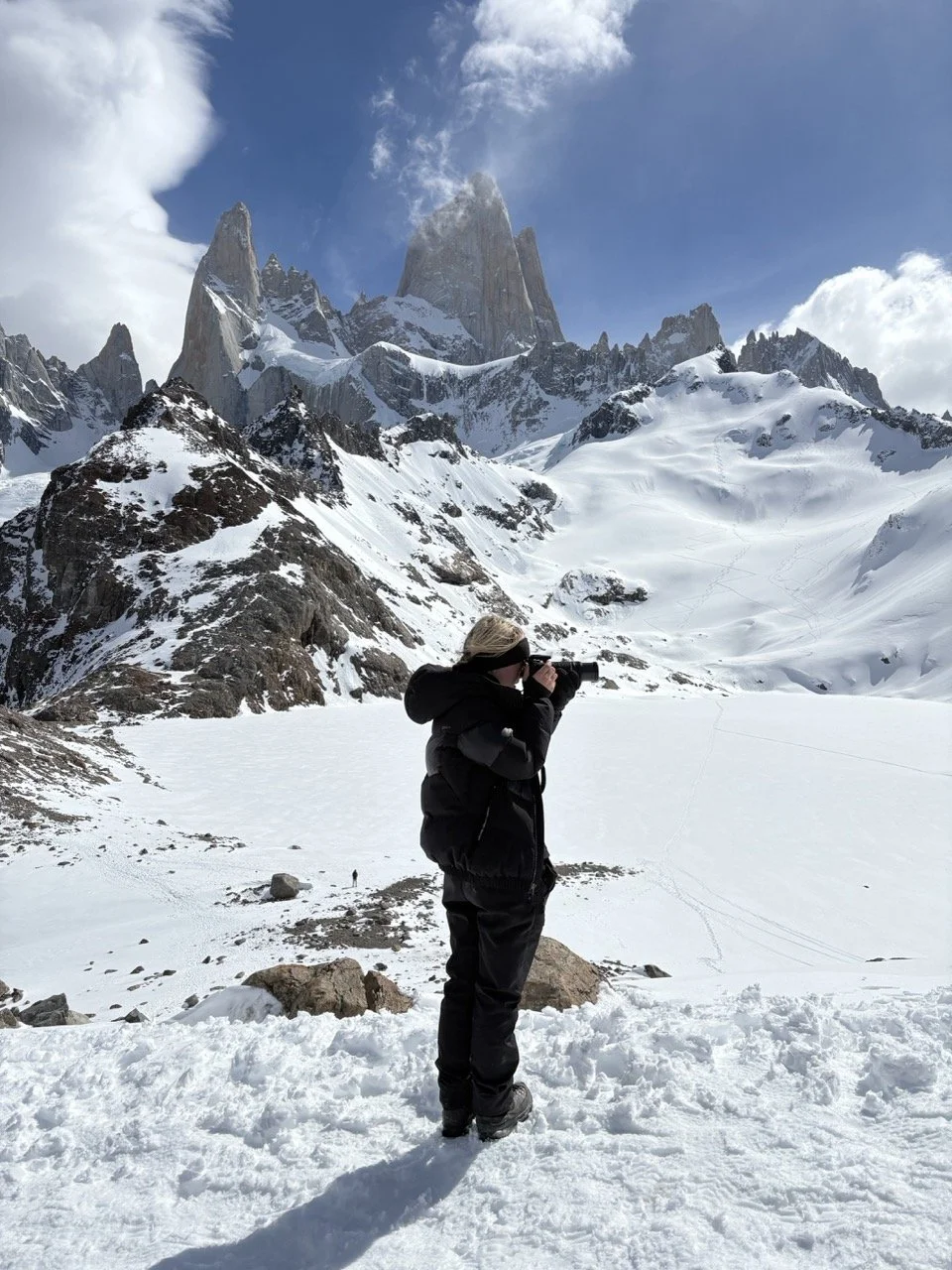 Person in black winter gear with hood and hat taking a photograph of snowy mountain peaks with clouds in the sky.