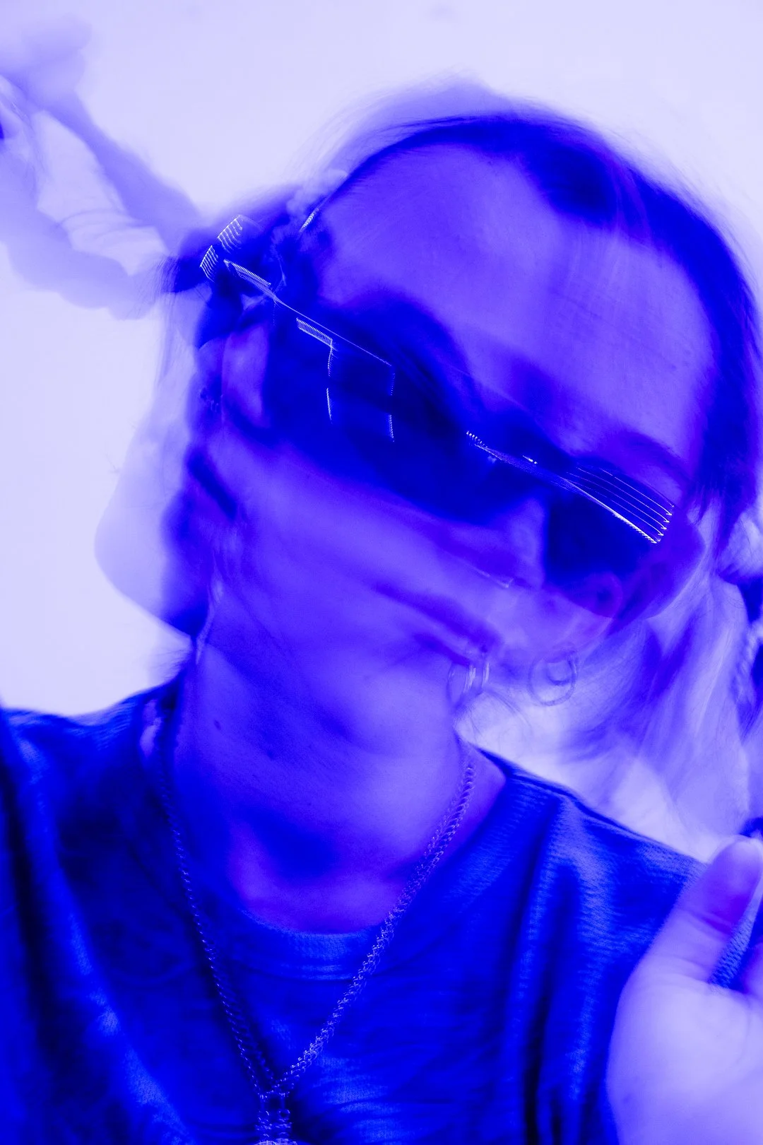 A person wearing glasses and a chain necklace, with a blue neon light seems to be capturing motion blur in the photograph.