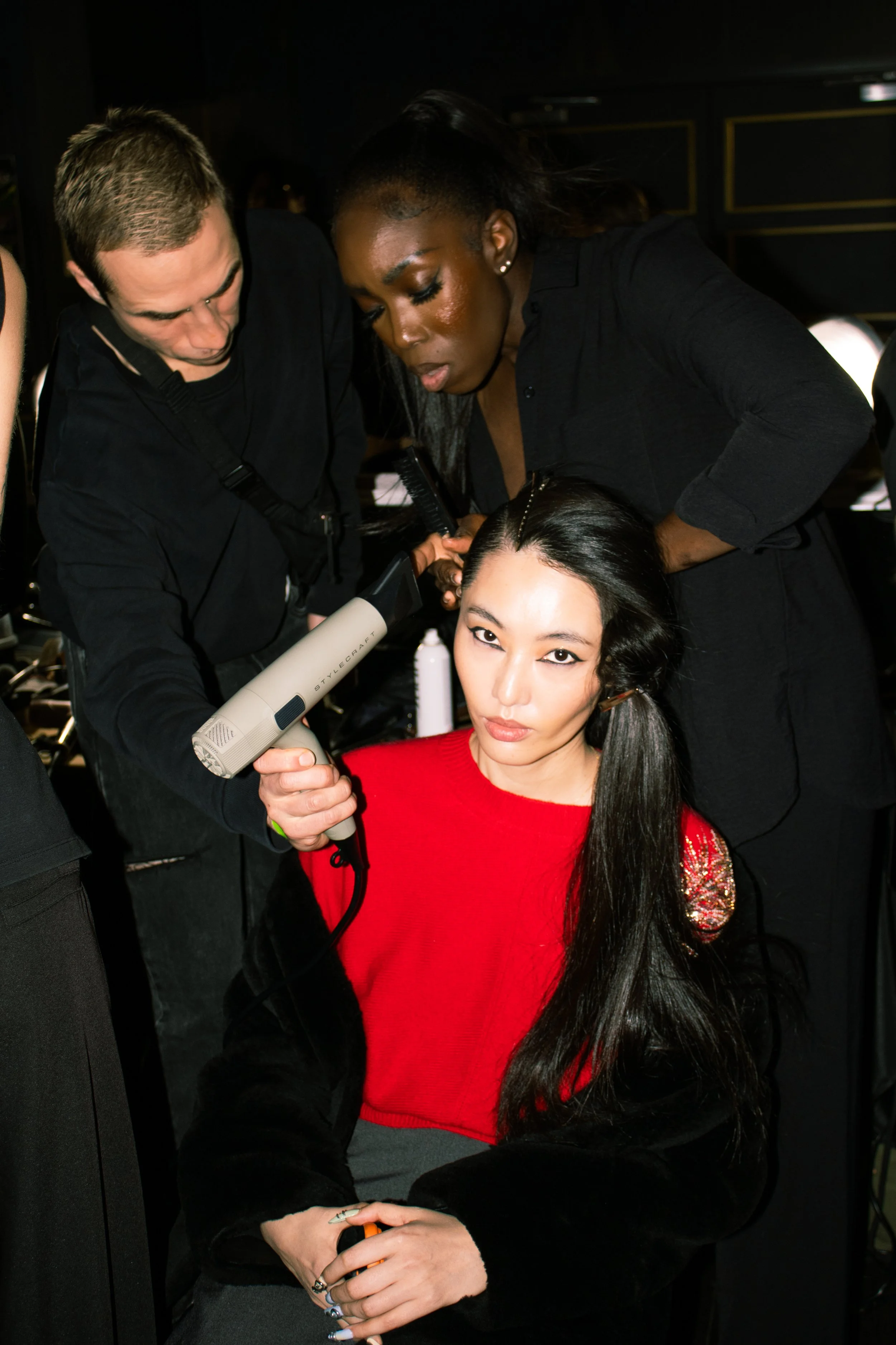 A woman with long black hair and a red top is sitting while two hairstylists are styling her hair at a salon or backstage setting.
