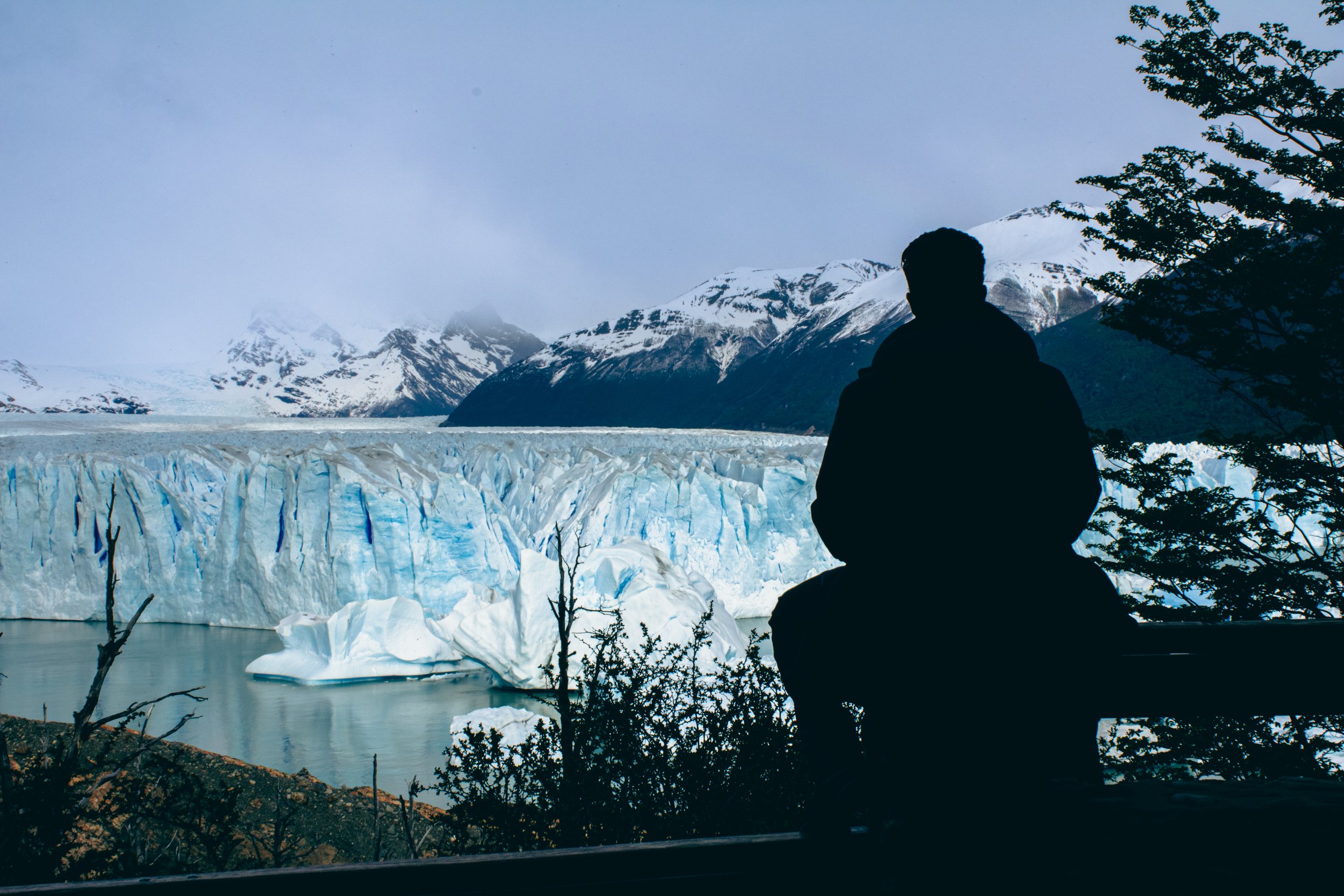 Silhouette of a person sitting on a bench overlooking a glacier and snow-capped mountains.