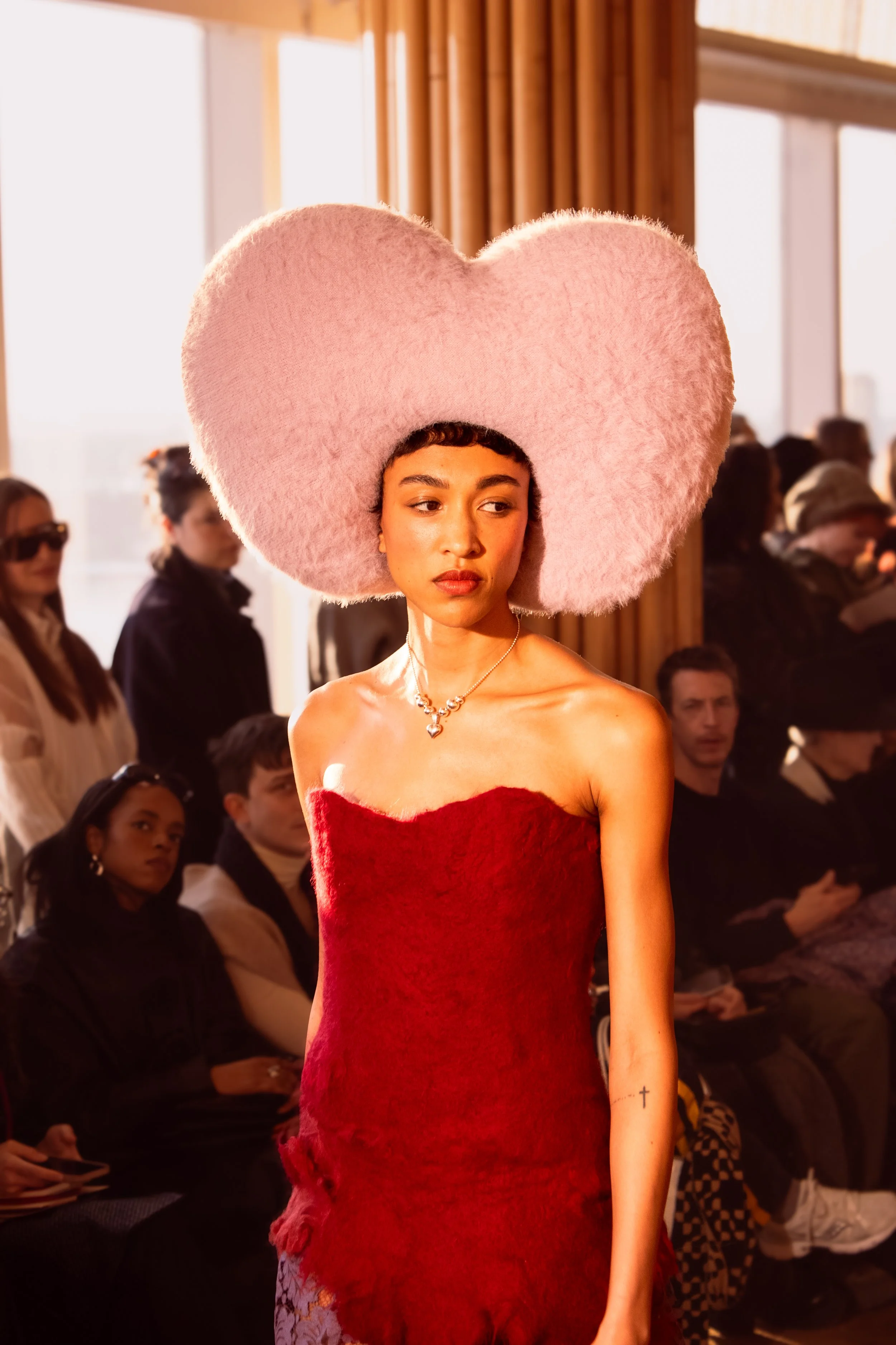 A young woman walking on a runway during a fashion show, wearing an oversized pink fluffy headpiece and a strapless red dress.