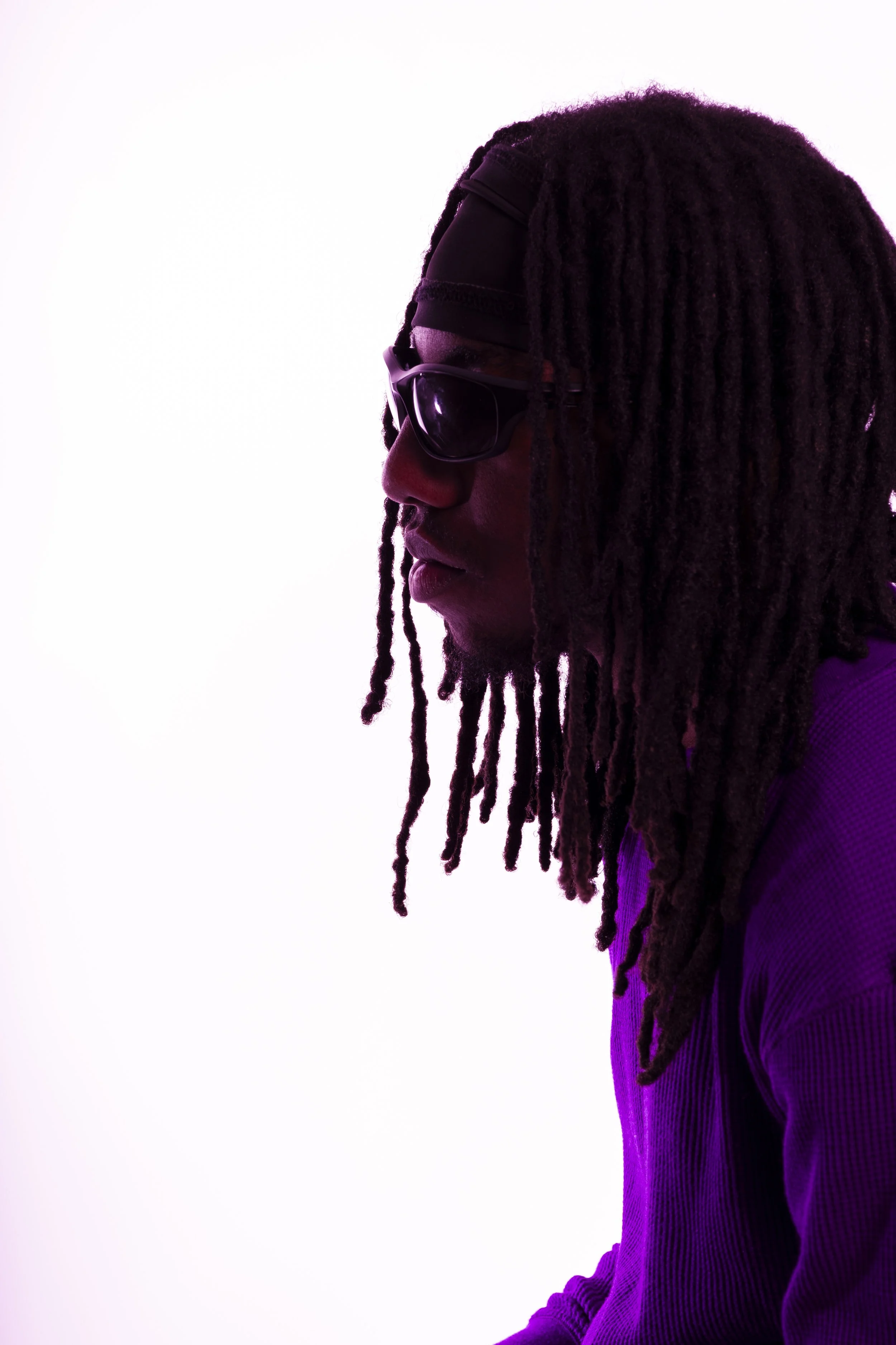 Profile of a man with long dreadlocks wearing sunglasses and a headband, illuminated with purple lighting against a white background.