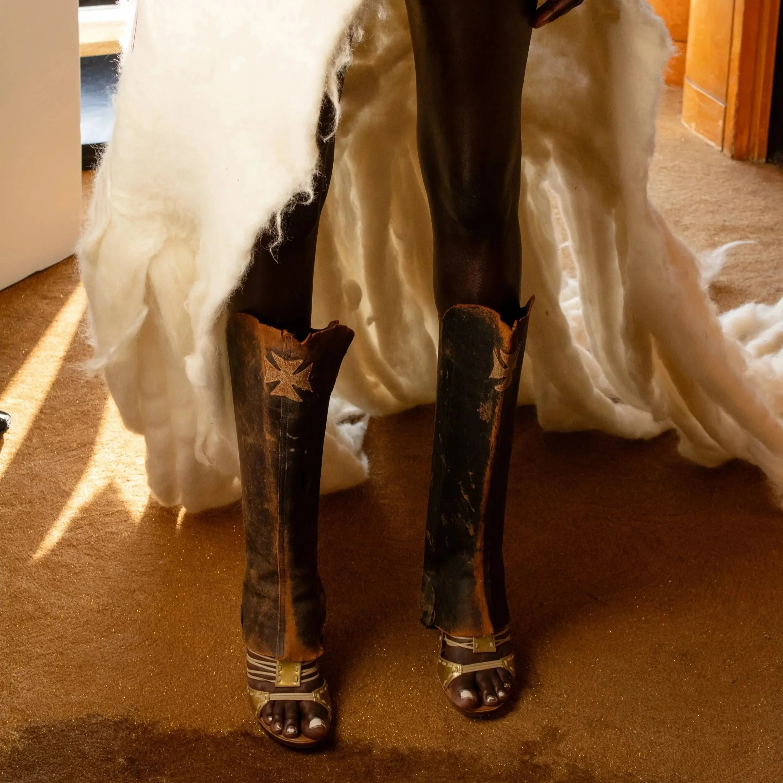 Close-up of the lower half of a person wearing leather chaps with star-shaped cutouts, tall cowboy boots, and a long, veiled dress, indoors with sunlight streaming on a carpeted floor.