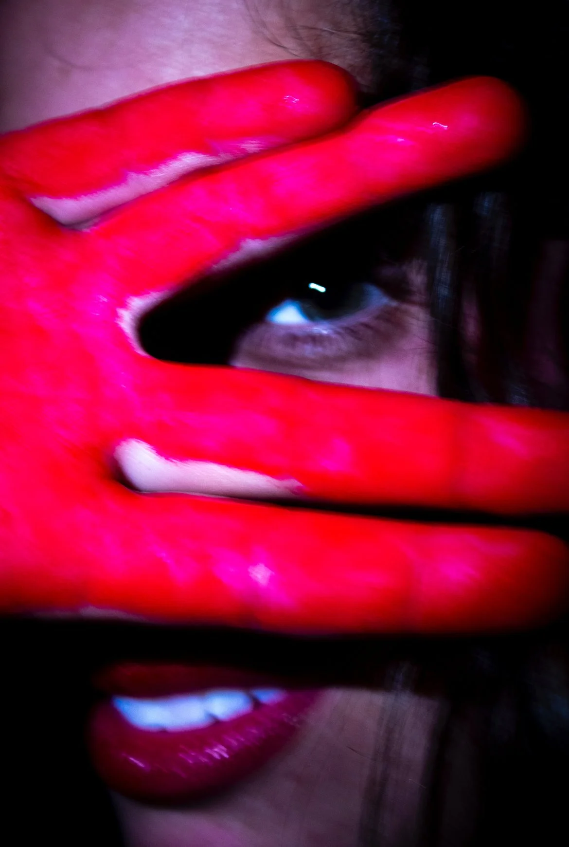 Close-up of a person with their face partly covered by their hand, which is painted bright red, with visible lips and eye behind the fingers.