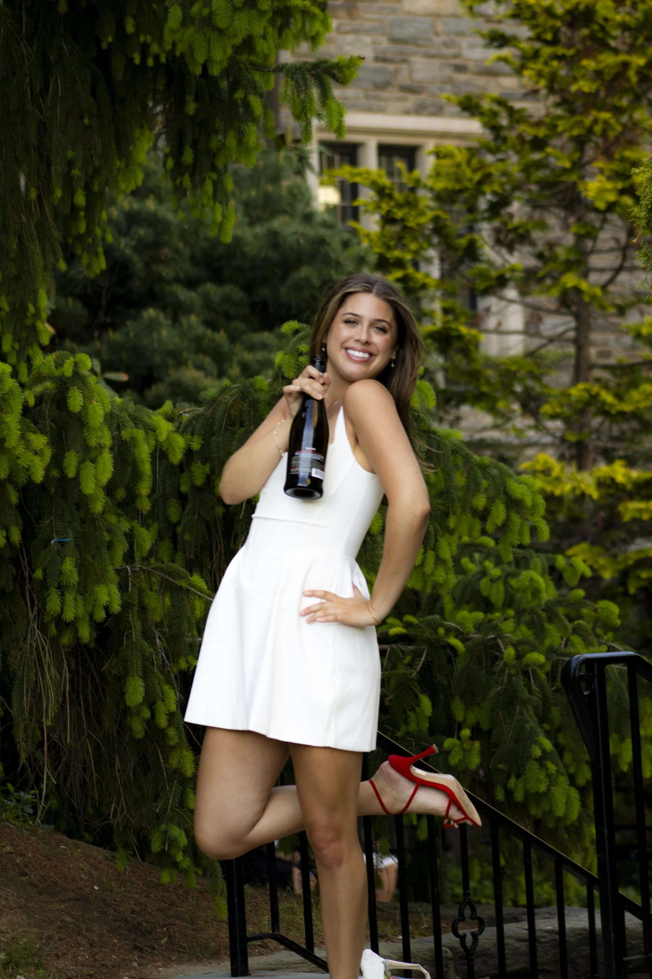 Woman in white dress smiling, holding a champagne bottle, outdoors surrounded by green trees and bushes, standing on steps with one foot raised, wearing red high heels.