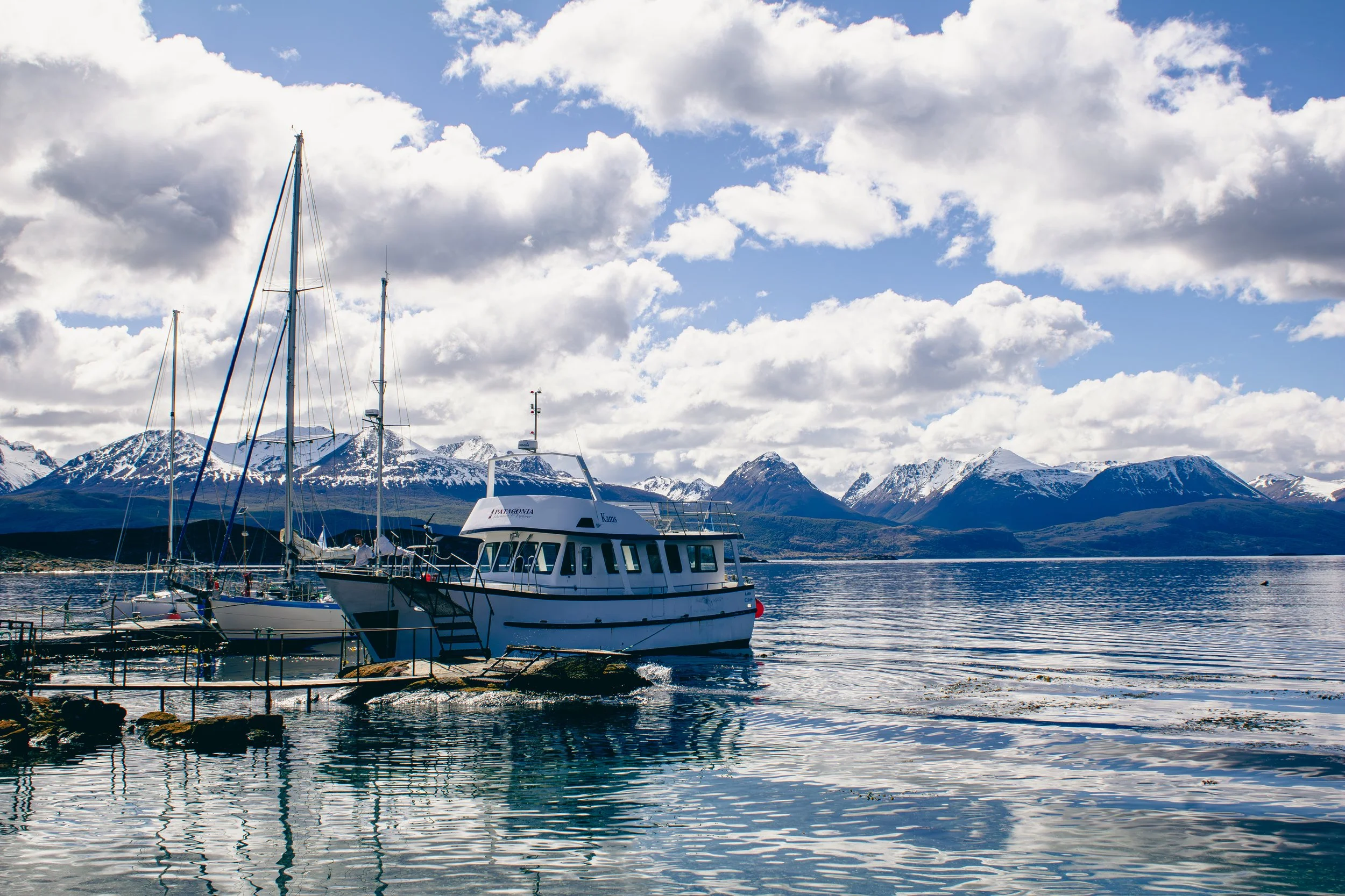 Boats docked on a calm lake with snow-capped mountains in the background under a partly cloudy sky.
