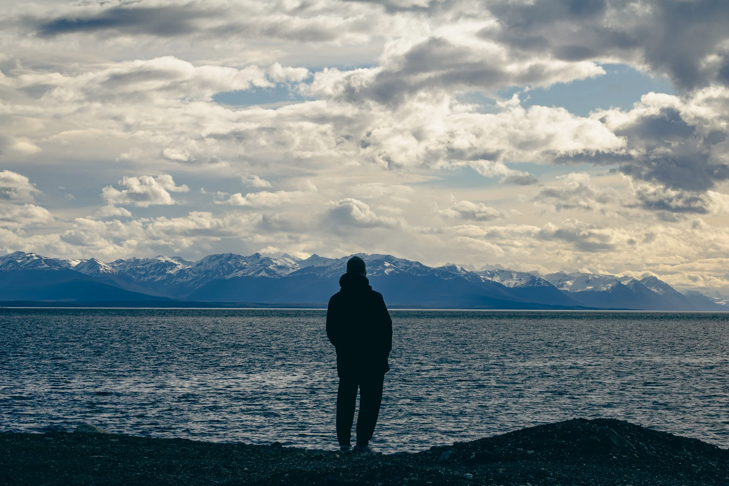 A person stands at the edge of a body of water, facing mountains with snow, under a cloudy sky.