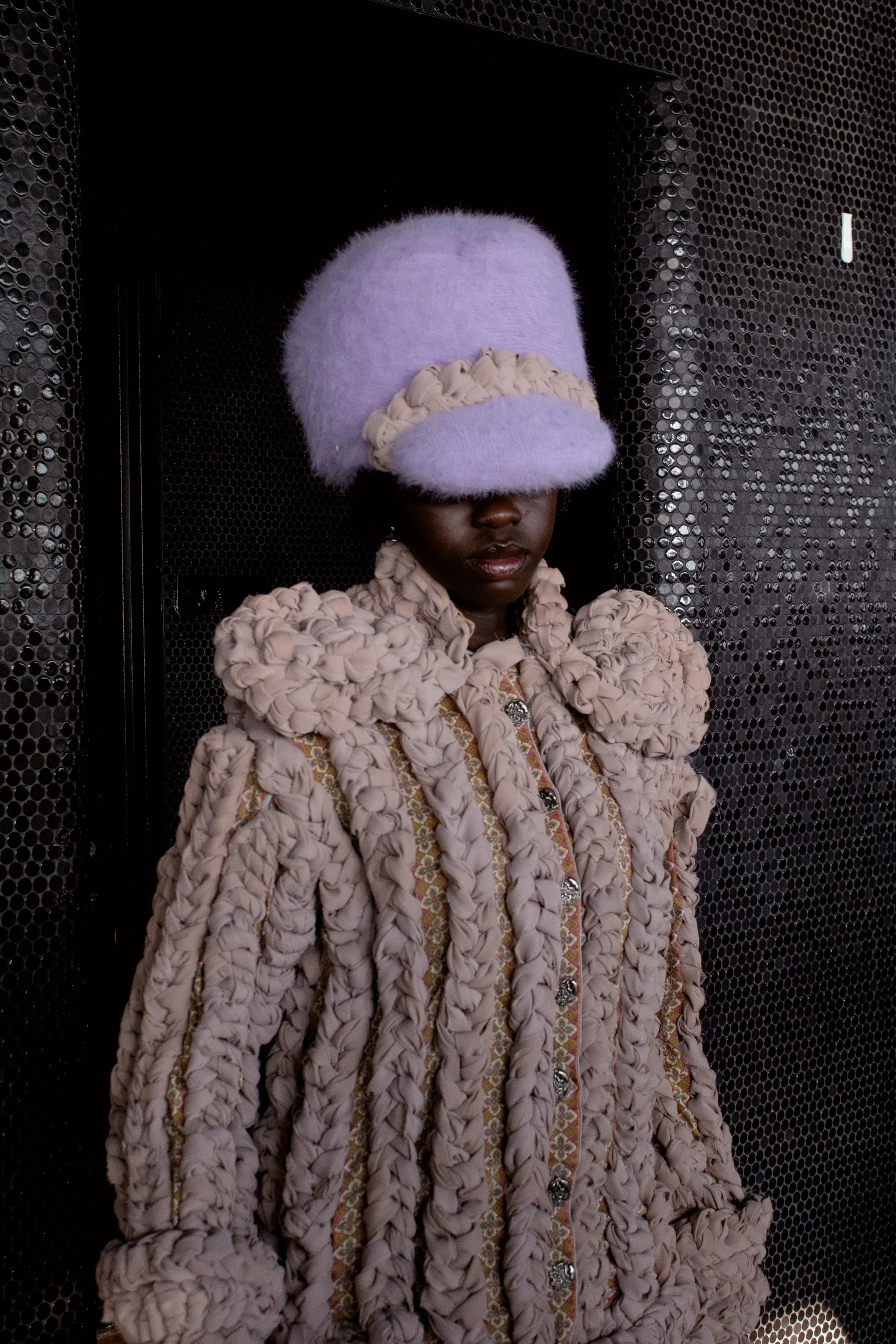 A person wearing an oversized lavender fluffy hat and a purple textured coat with puffed details, standing against a dark perforated metal background.