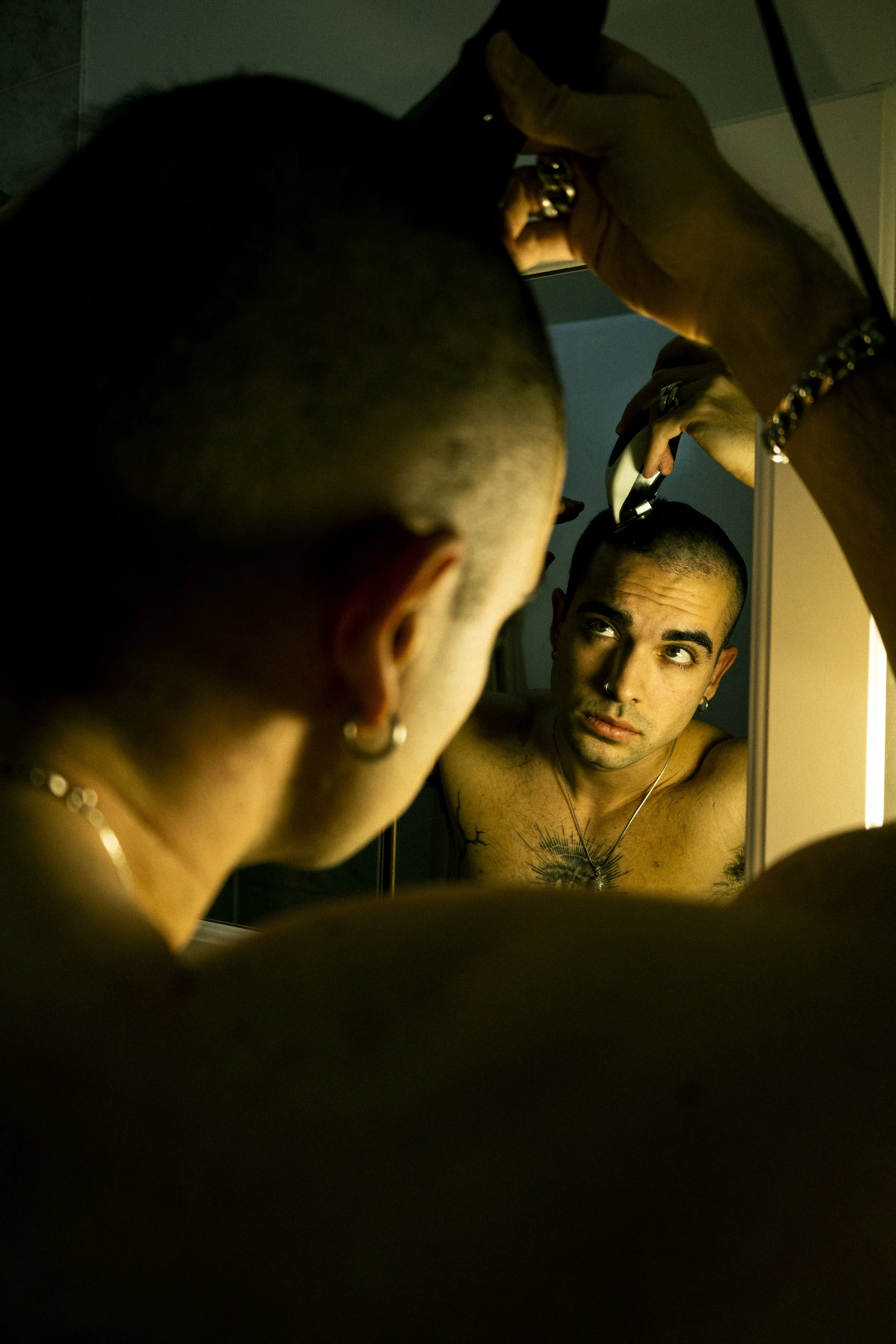 A man with a shaved head looking at himself in a mirror while getting a haircut or grooming, with hair clippers in hand, in a dimly lit room.