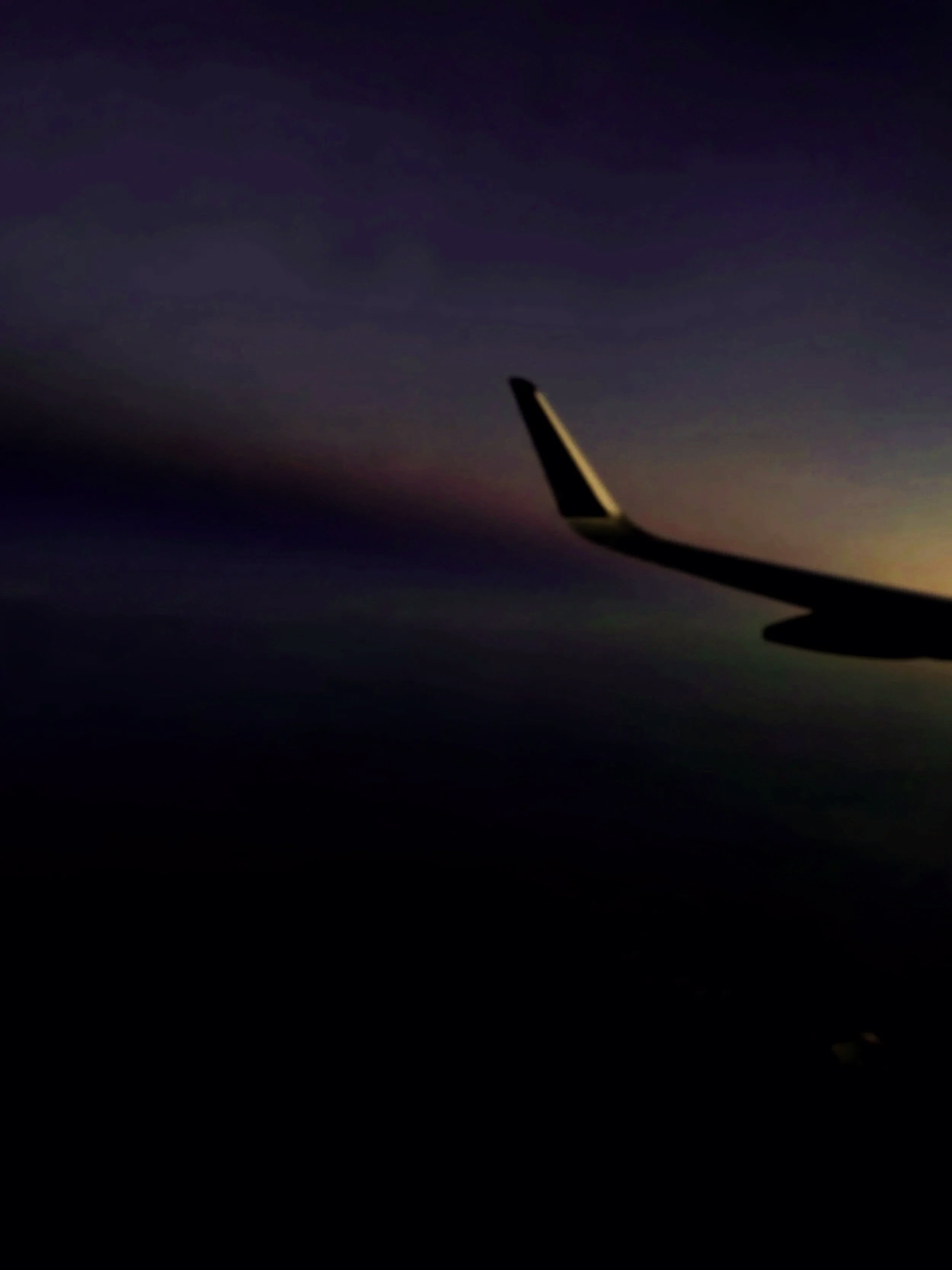 Photo of airplane wingtip against a dark, colorful sky during sunset or sunrise, viewed from the window of an airplane.