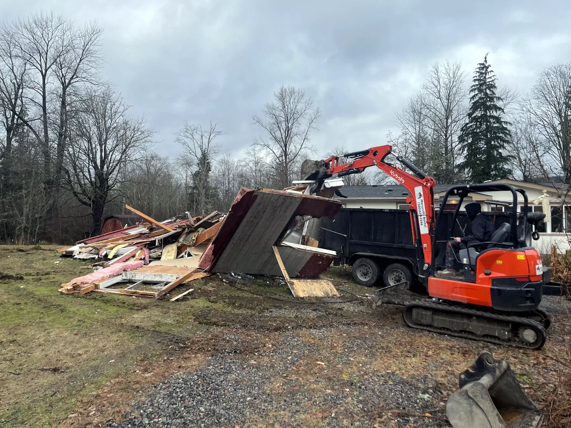 A small excavator with an orange body and black tracks demolishing a pile of debris including wood, insulation, and roofing materials outdoors under a cloudy sky.