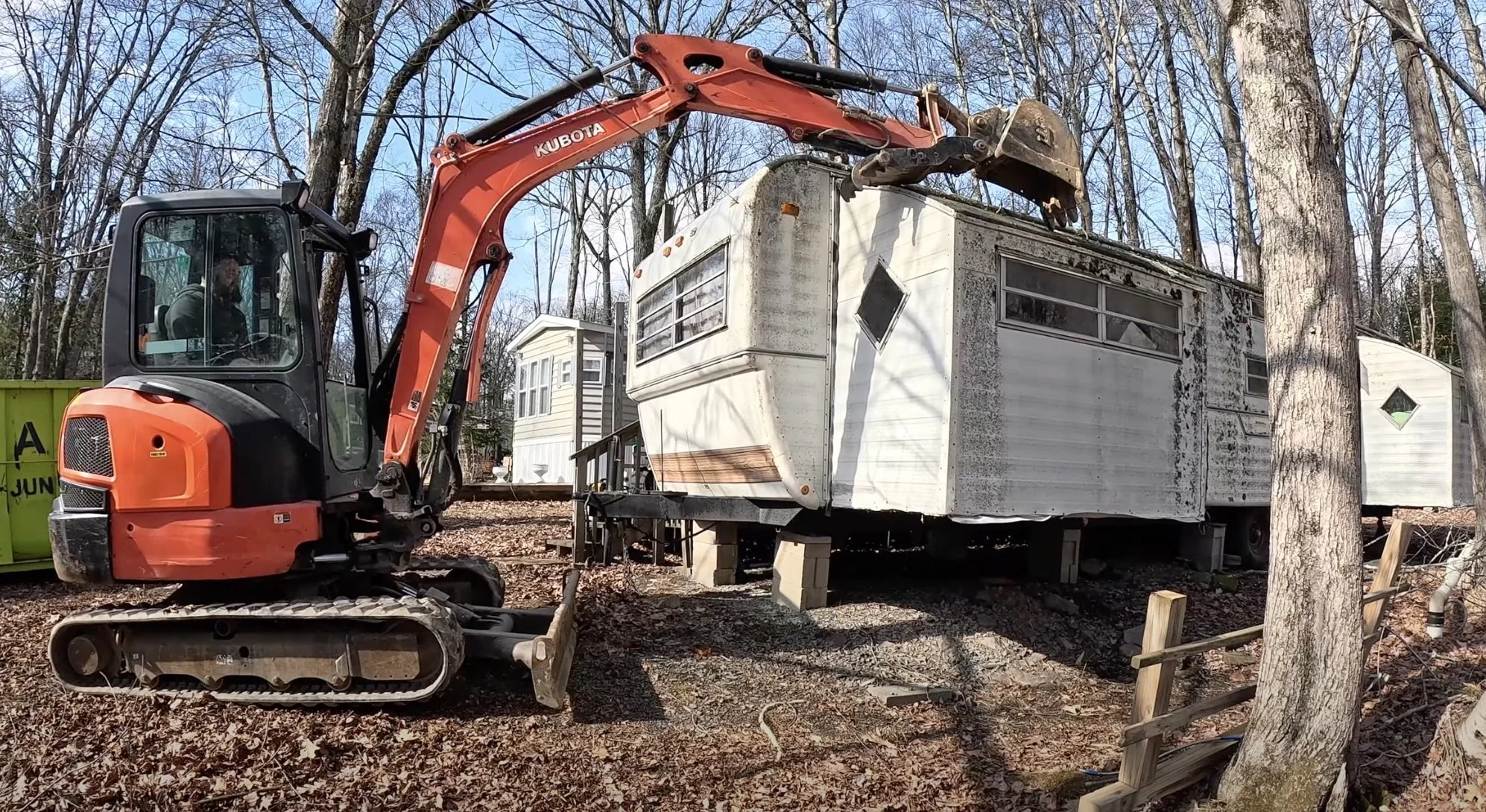 A Kubota excavator with an orange arm demolishing or moving an old, weathered mobile home in a wooded area.