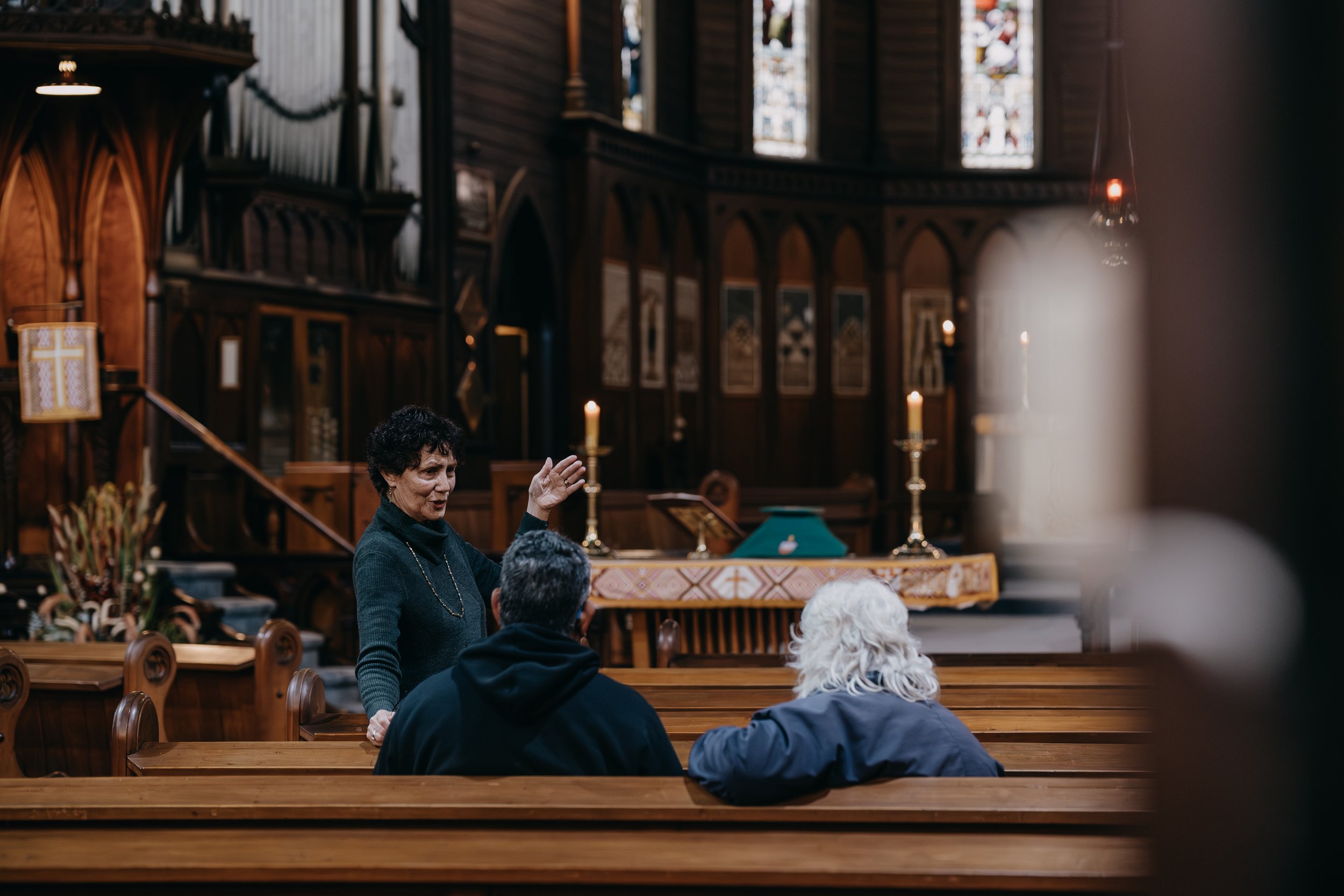 A woman speaking or singing in a church with stained glass windows, candles, and altar decor, while people sit on benches listening.