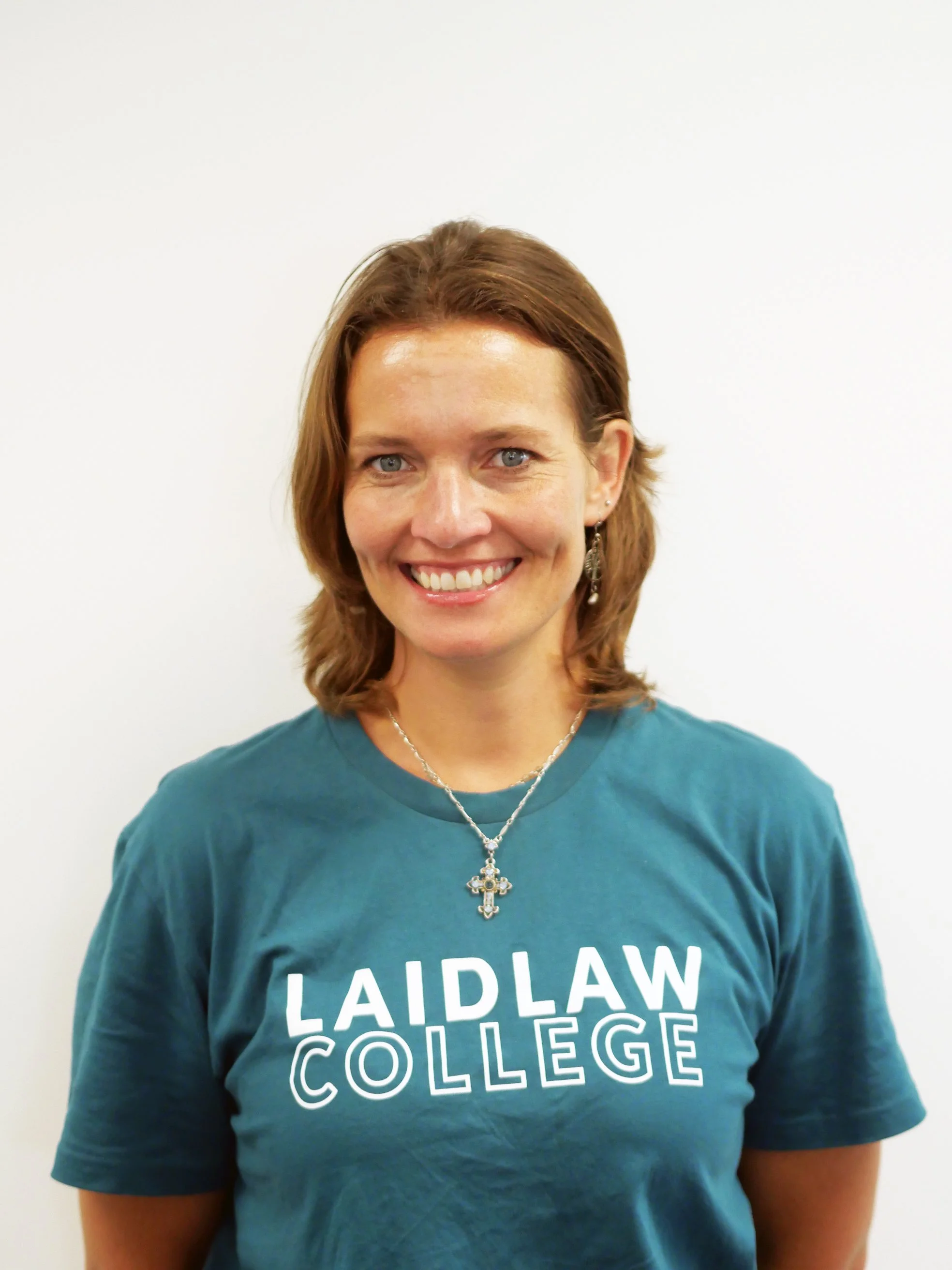 A photo of Louise Wernich with shoulder-length light brown hair, blue eyes, smiling, wearing a teal T-shirt with the words 'LAIDLAW COLLEGE' printed on it, and a silver necklace with a cross pendant, standing against a plain white background.