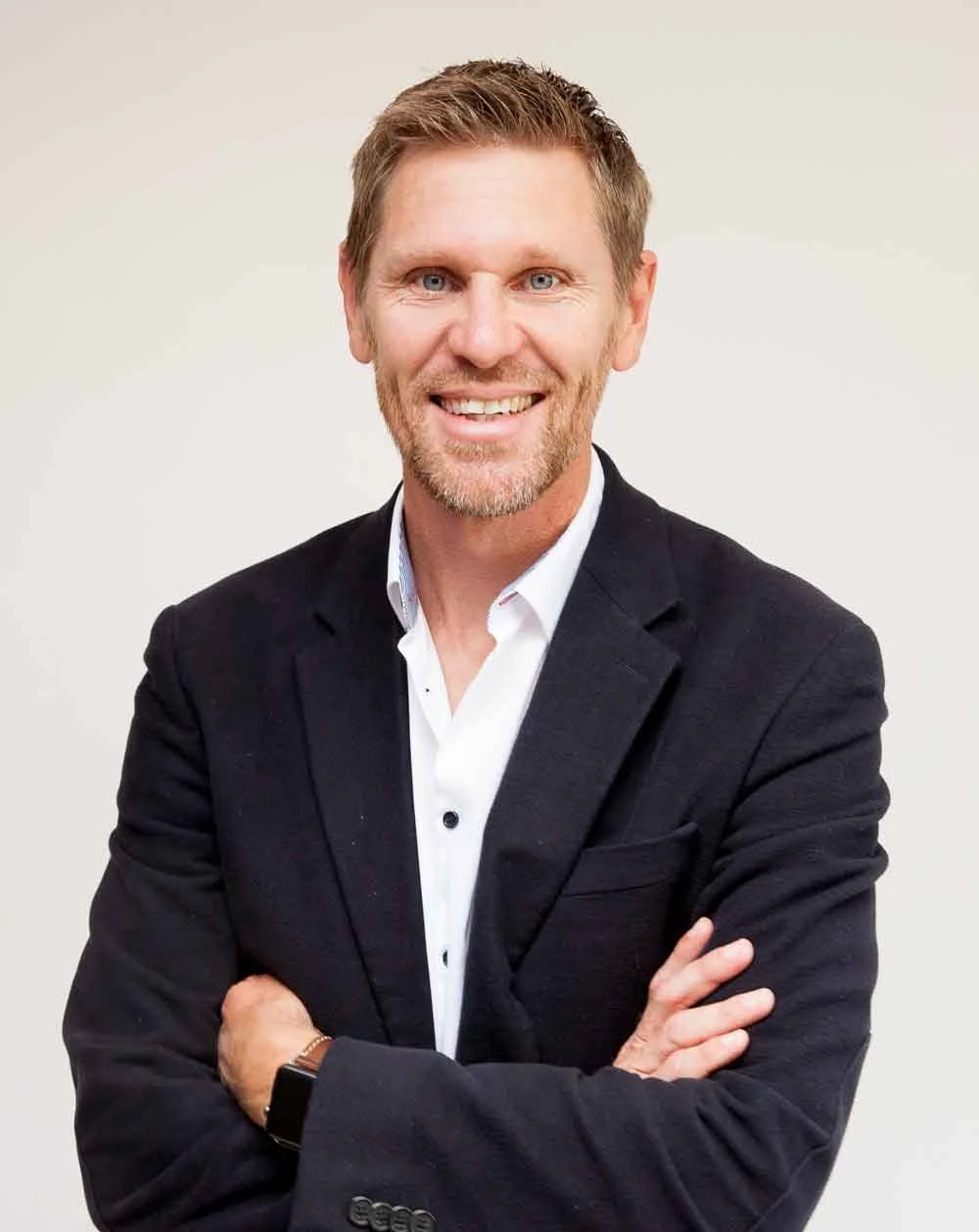 Jonathan, a portrait of a smiling man in a black blazer with crossed arms, white shirt, and a watch, standing against a plain light background.
