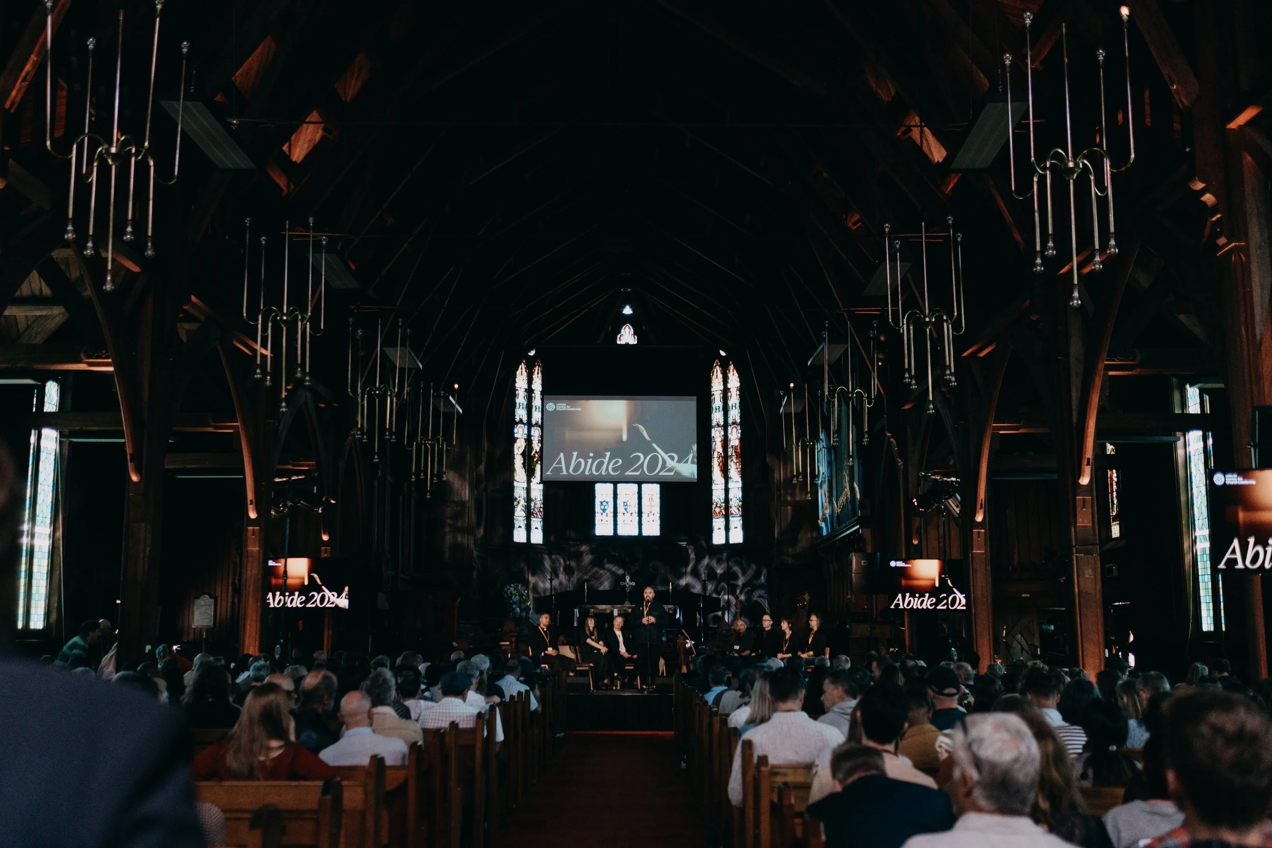 Interior of a dark wooden church or hall filled with an audience seated in rows, facing a stage with a large screen displaying the words 'Abide. The ceiling has wooden beams and hanging metal chandeliers. There are stained glass windows along the sides of the hall.