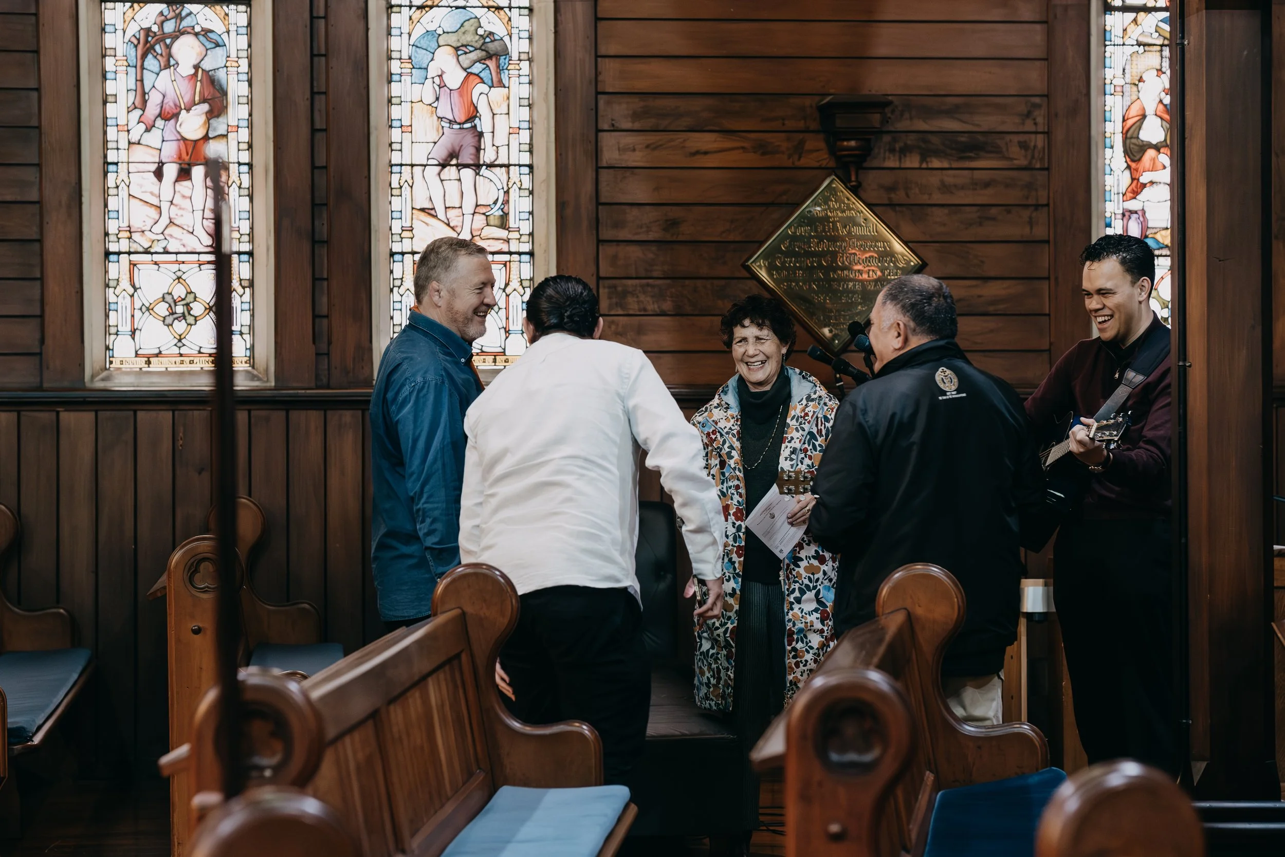 A group of people inside a church with wooden walls and stained glass windows, some sitting and some standing, engaged in conversation and singing.