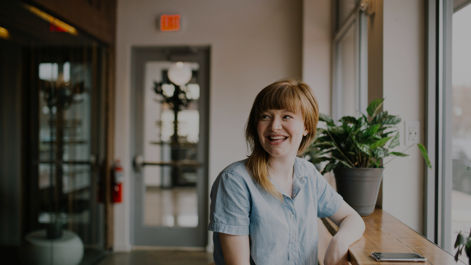 A woman with red hair, smiling, sitting by a window with a potted plant and a smartphone on the table.
