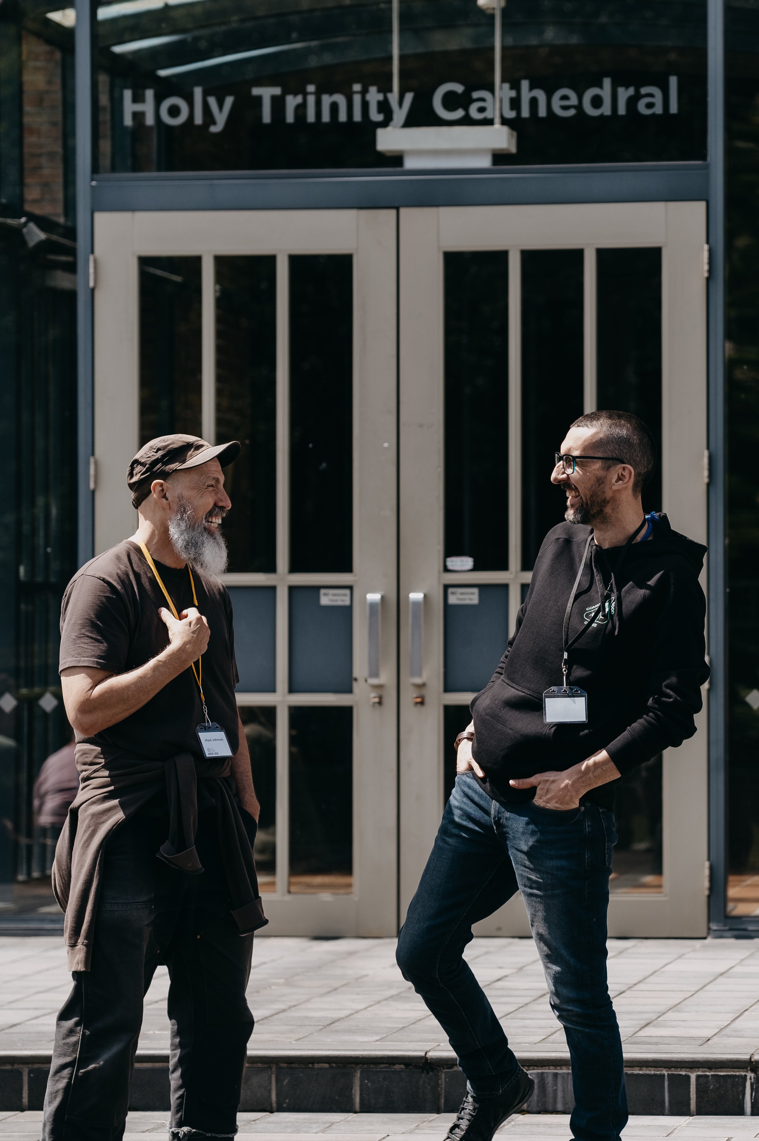 Two men with lanyards and badges talking outside in front of Holy Trinity Cathedral, smiling and laughing at each other.