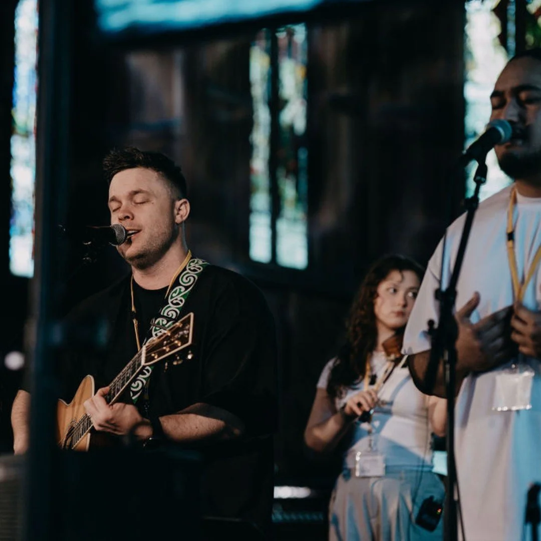 A man with closed eyes singing into a microphone while playing an acoustic guitar, with two other people, a woman holding a tambourine and a man with a microphone, in a dimly lit setting with stained glass windows in the background.