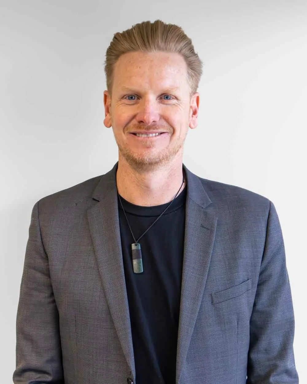 Clint Ussher, a smiling man with light skin, short blonde hair, wears a dark blazer, black shirt, and a necklace with a rectangular pendant.