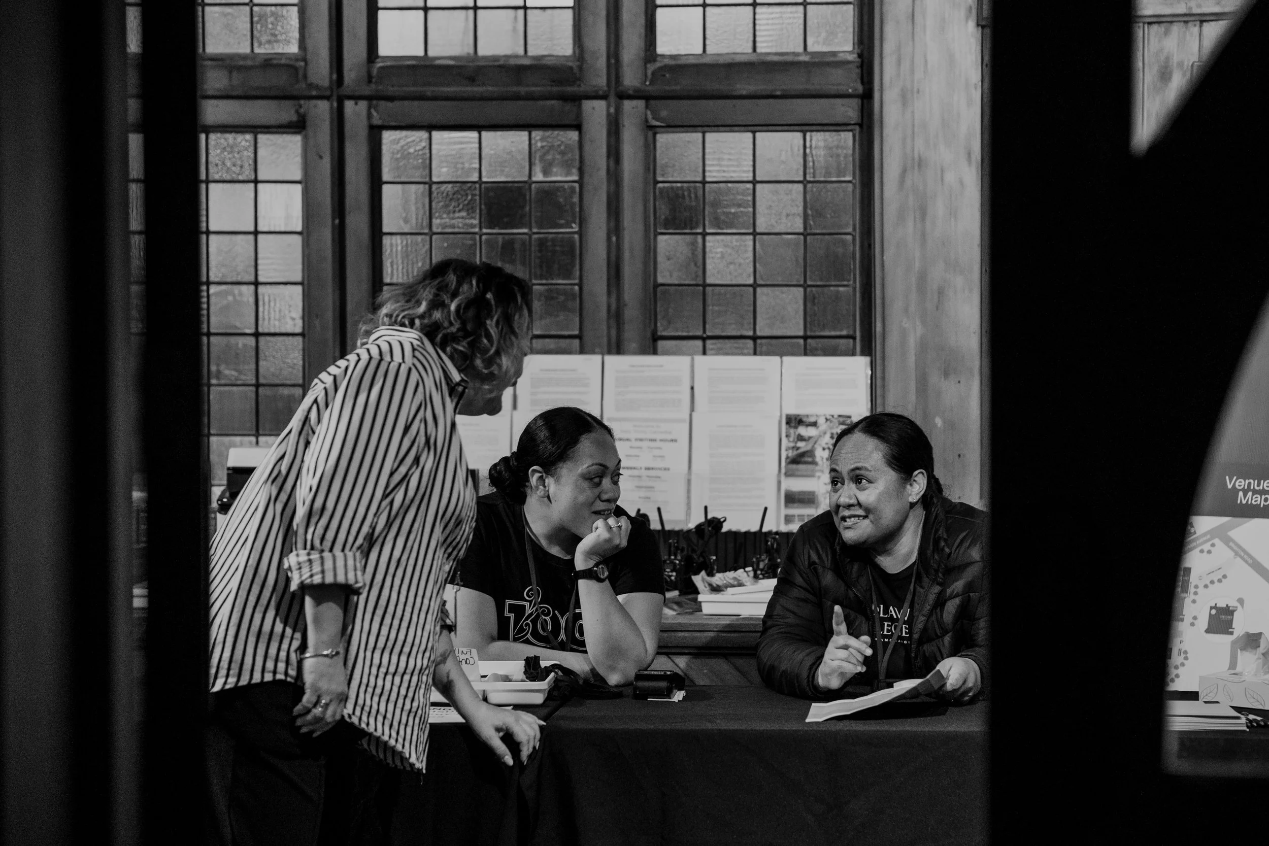 Three women engaged in a conversation at a registration or information booth inside a building with large, grid-patterned windows.