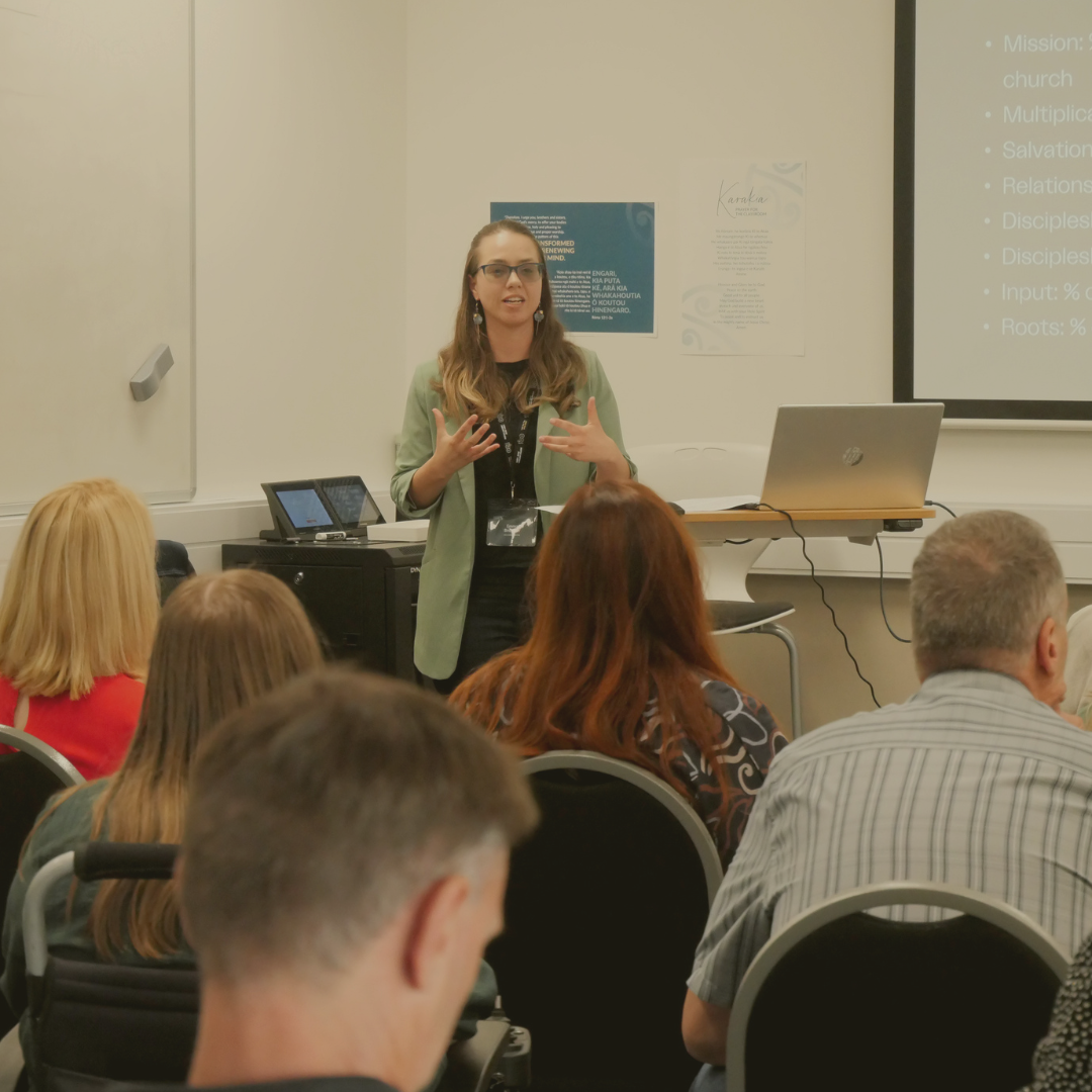 A woman with glasses and long hair, wearing a black top and green blazer, is giving a presentation in a classroom or conference room. She stands in front of a screen displaying a slide with text, and an audience is seated facing her. The room has white walls, and there are several posters on the wall behind her.