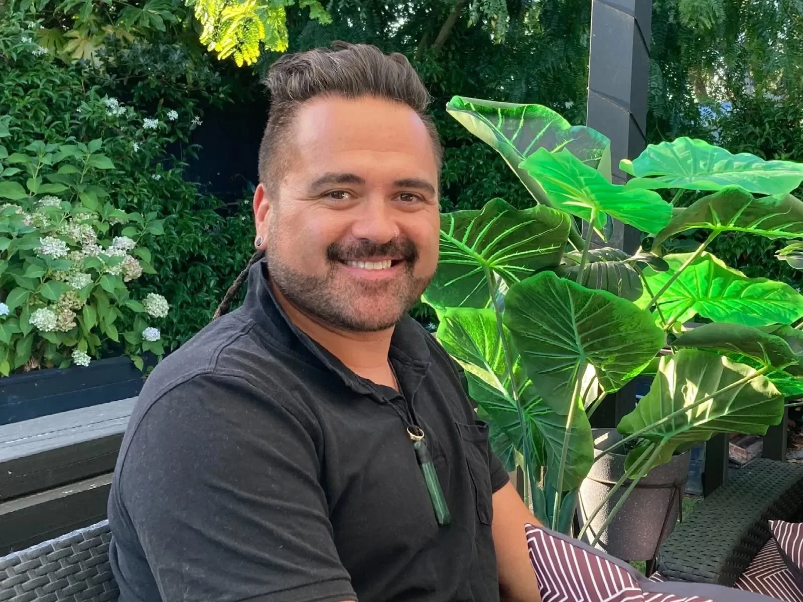 A man with brown hair and a beard, smiling, sitting outdoors surrounded by green plants, including large leafy plants and smaller flowering bushes.